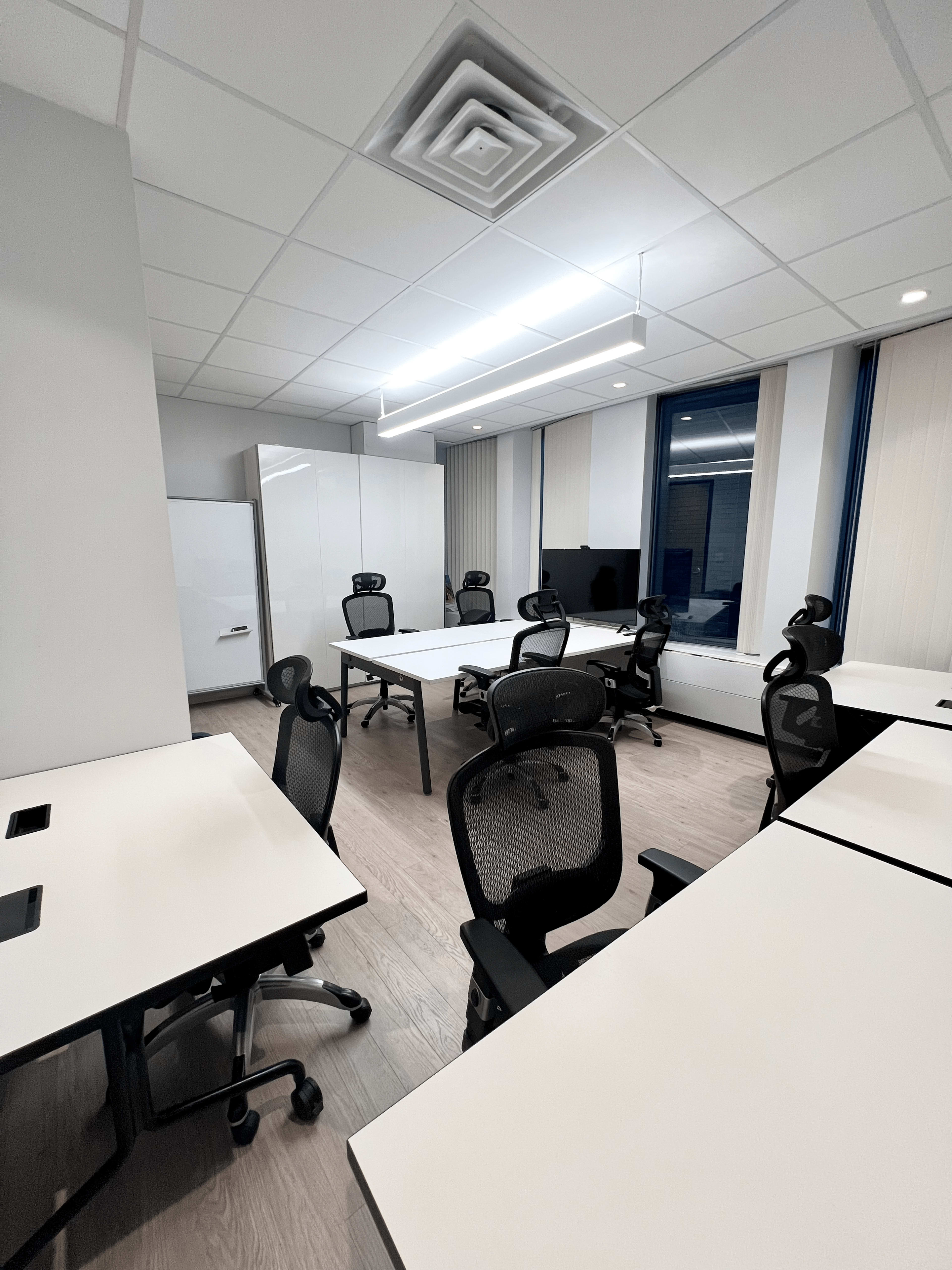 A modern office space with several white tables and ergonomic chairs arranged around the room.