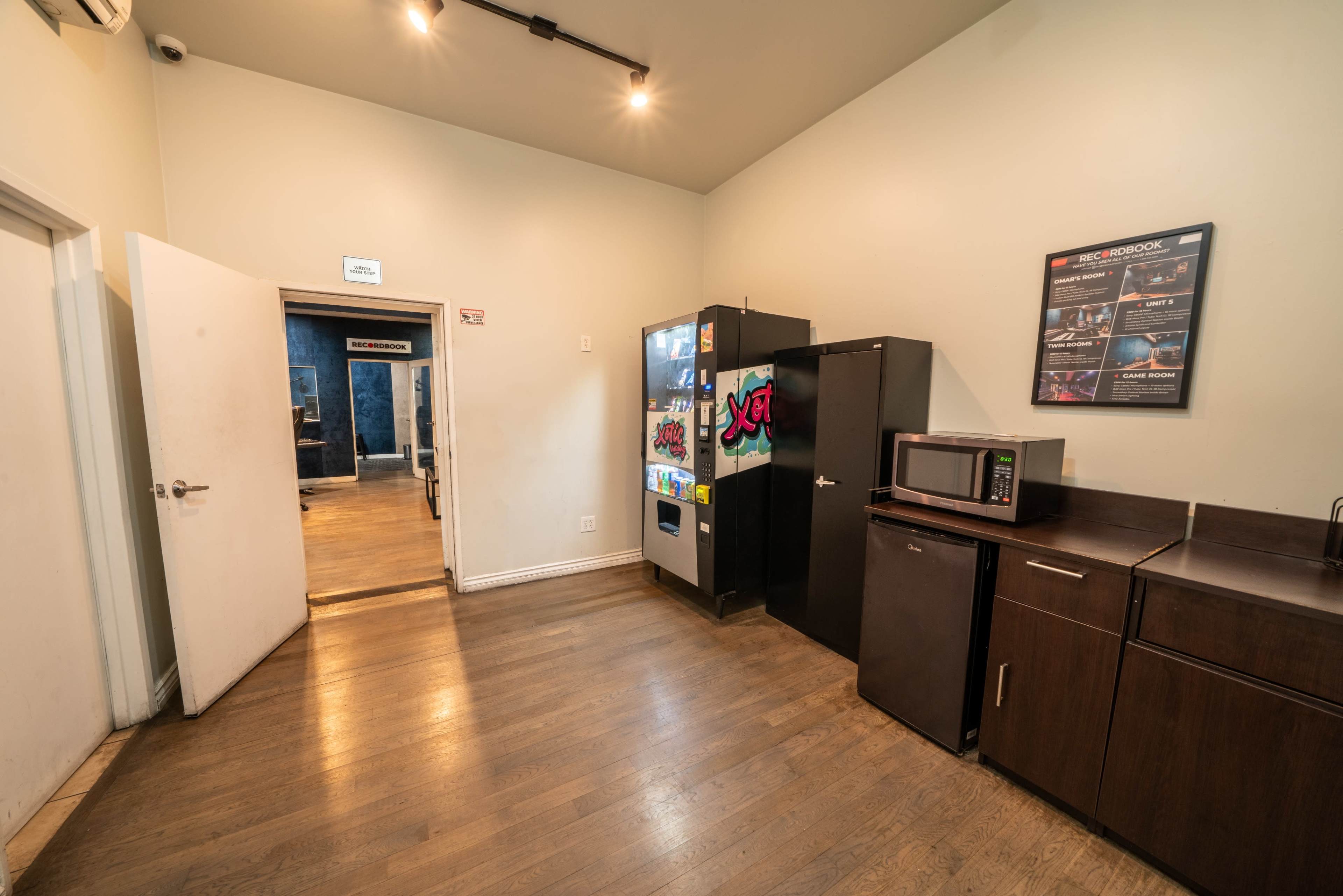 The image shows a small room with a microwave, a black refrigerator, and a vending machine against one wall, leading to another room through an open doorway.