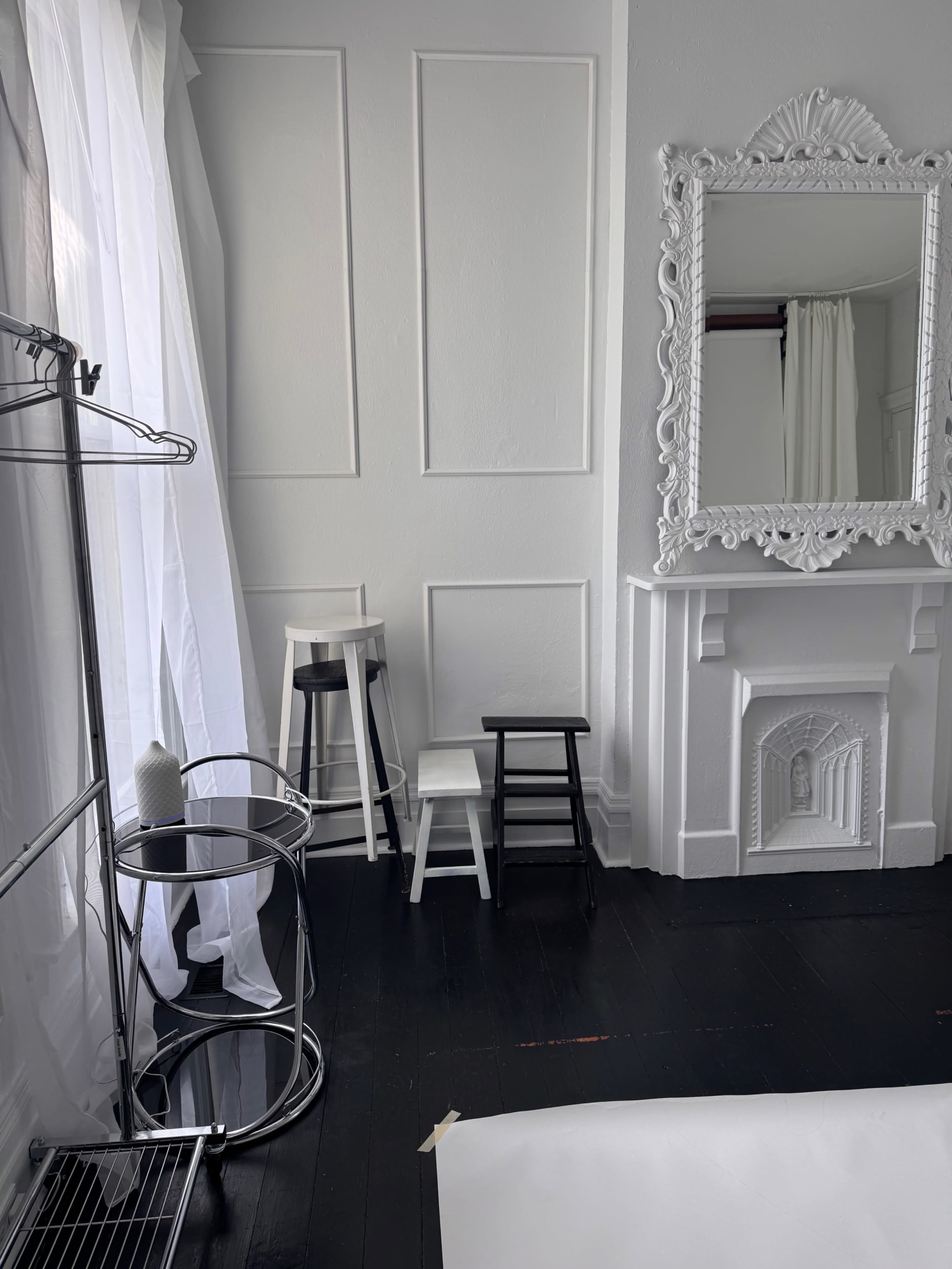The image shows a bright room with white walls, a large ornate mirror, three stools in different colors, a clothing rack, and a round table.