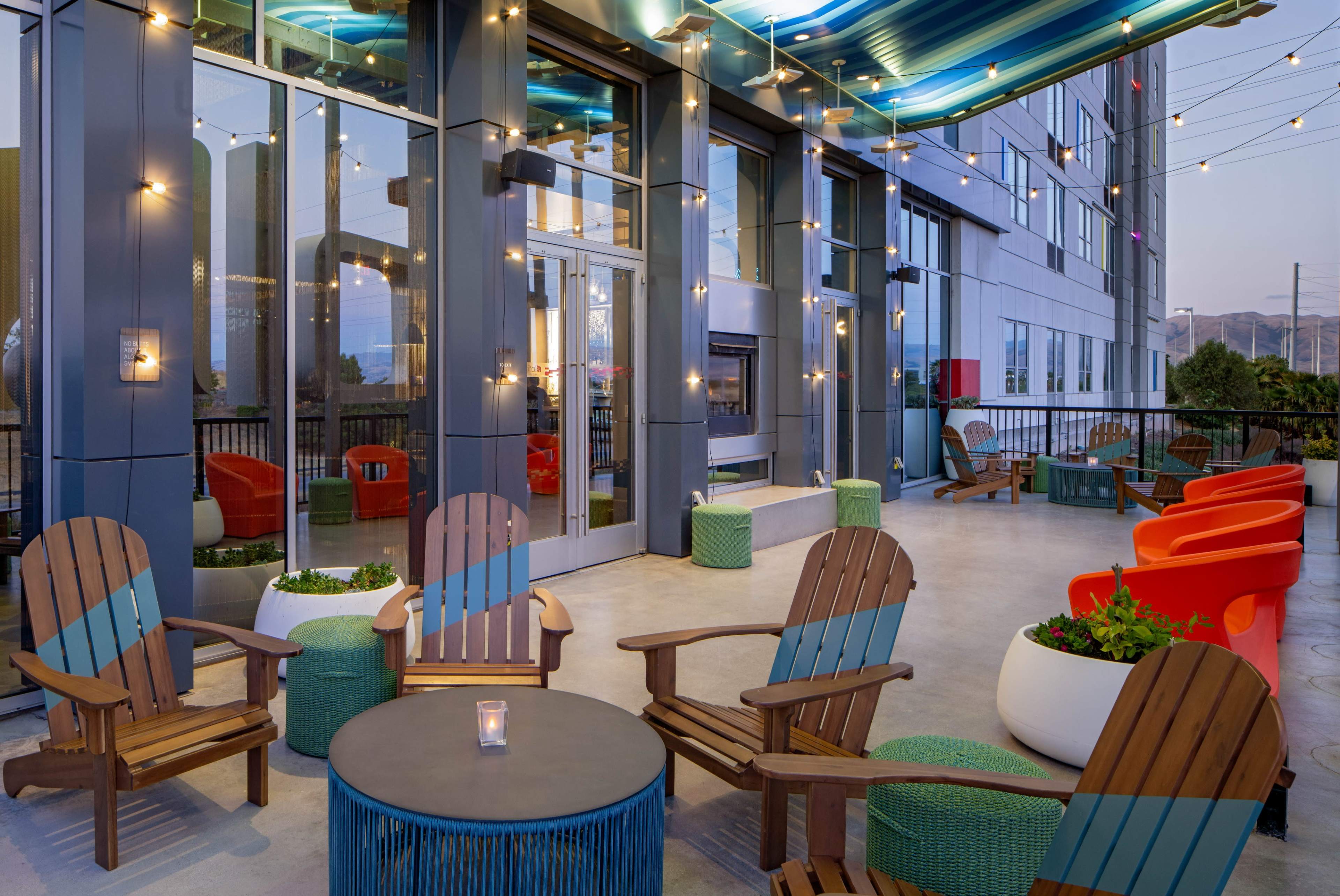 A modern outdoor seating area with colorful chairs, wooden lounge chairs, and potted plants under string lights.