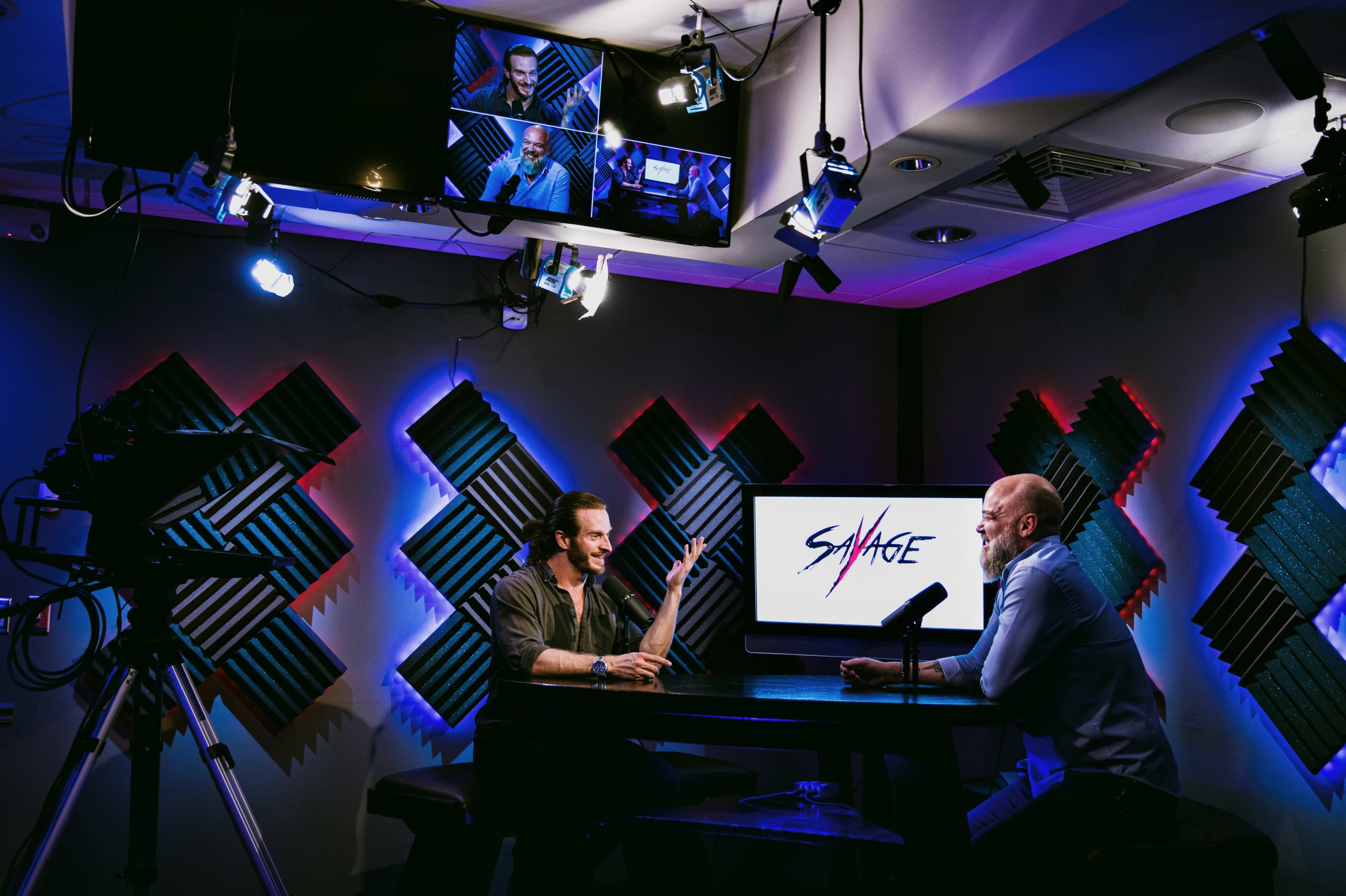 Two men are seated at a table in a studio, engaging in a conversation, with a screen displaying the word "SAVAGE" in the background and studio lights illuminating the scene.