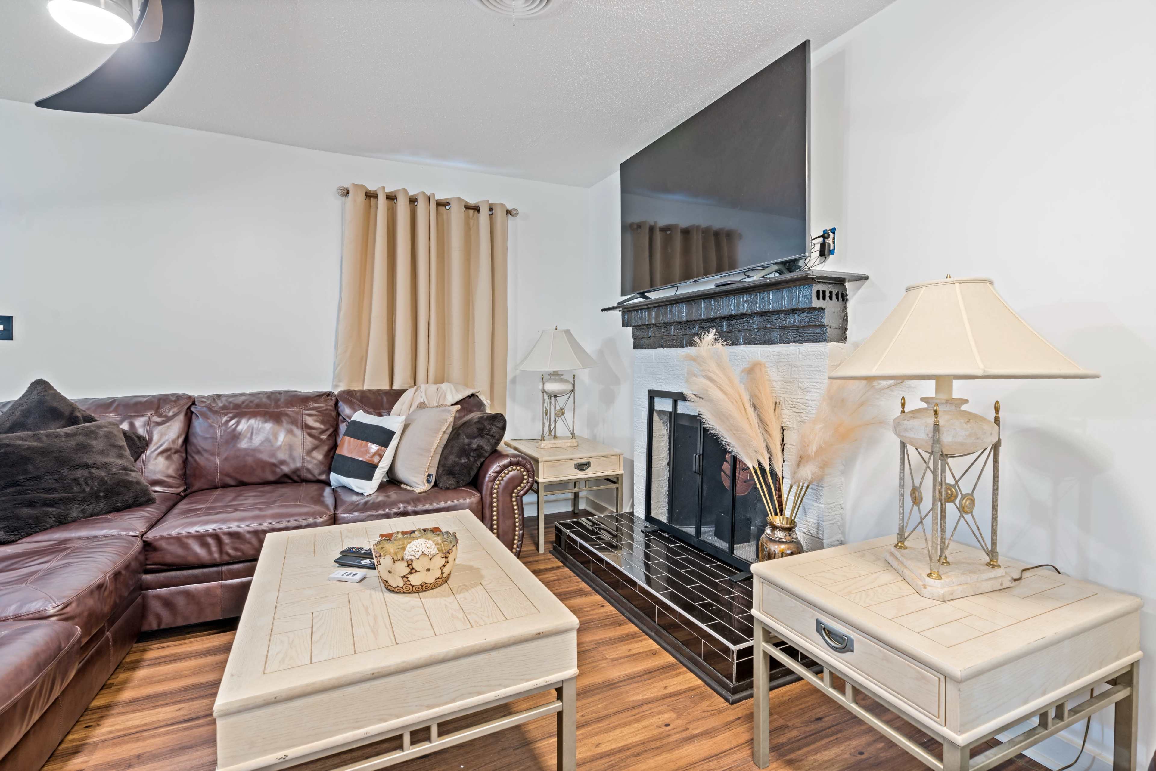The image shows a living room featuring a brown leather sofa, a coffee table with decorative items, and a fireplace surrounded by a black mantel, all set against a backdrop of beige curtains and wood flooring.