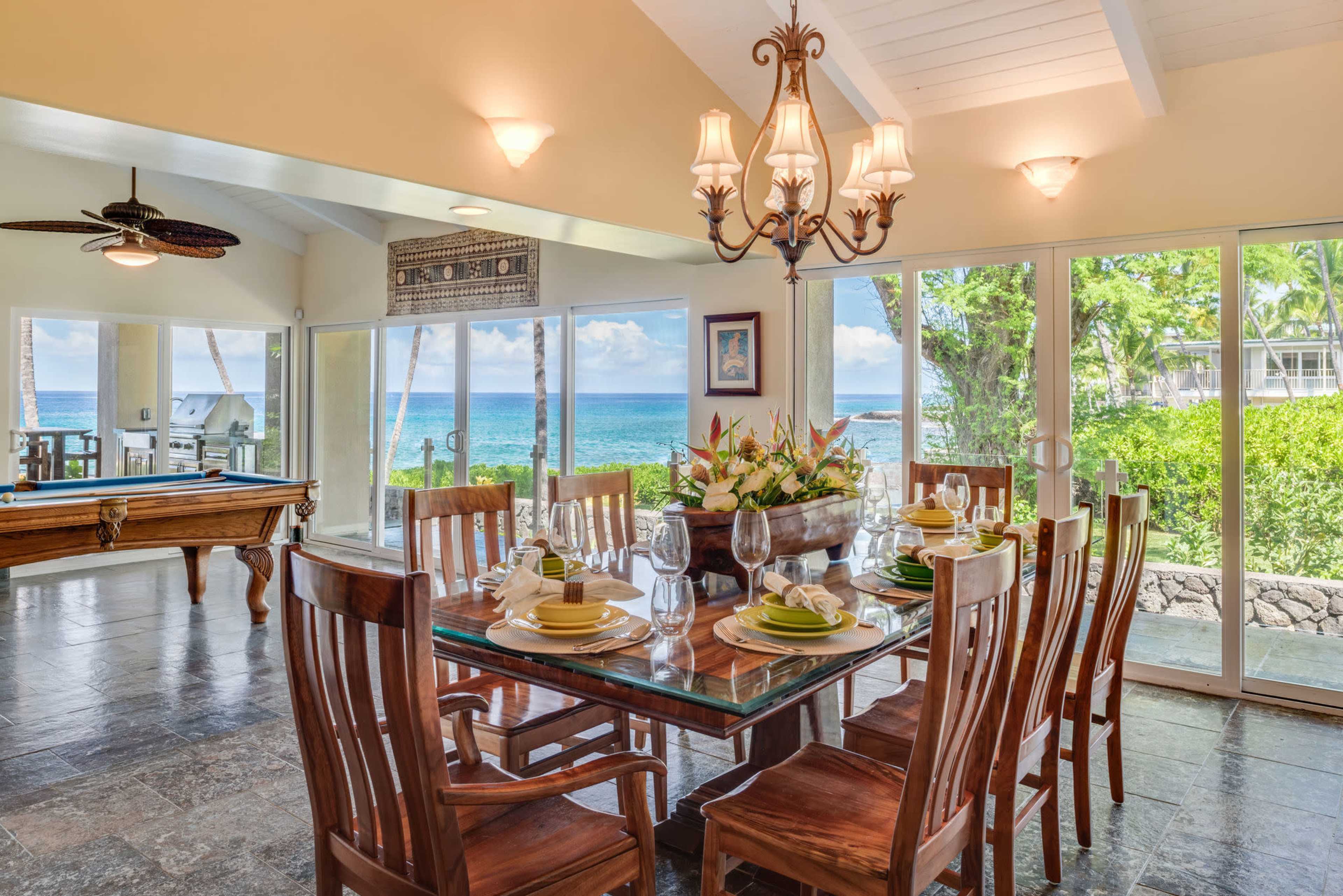 A dining area with a large table set for eight overlooks a view of the ocean through expansive glass doors.