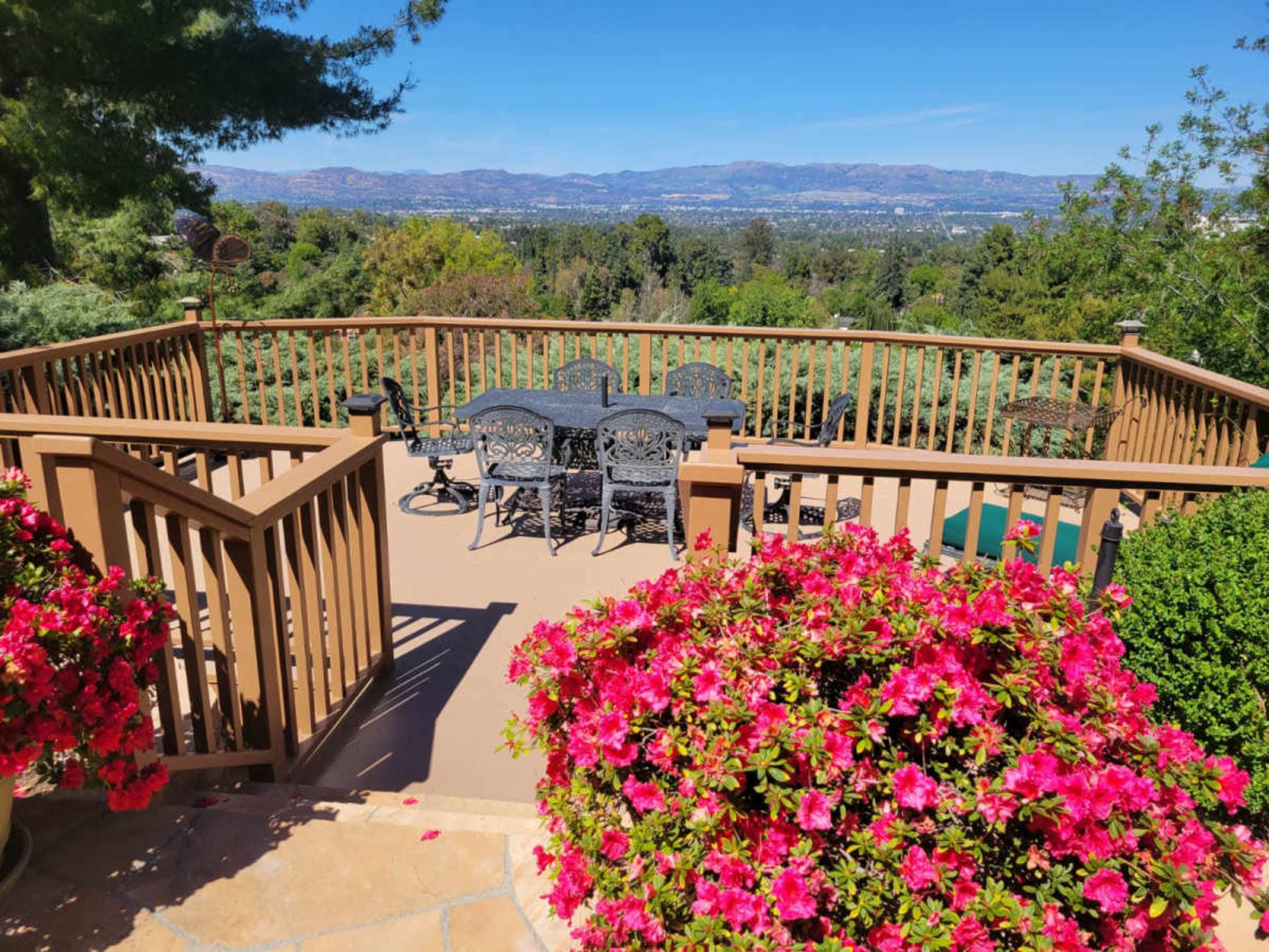 A wooden deck with a table and chairs overlooks a scenic valley filled with greenery and distant mountains, framed by blooming pink flowers.
