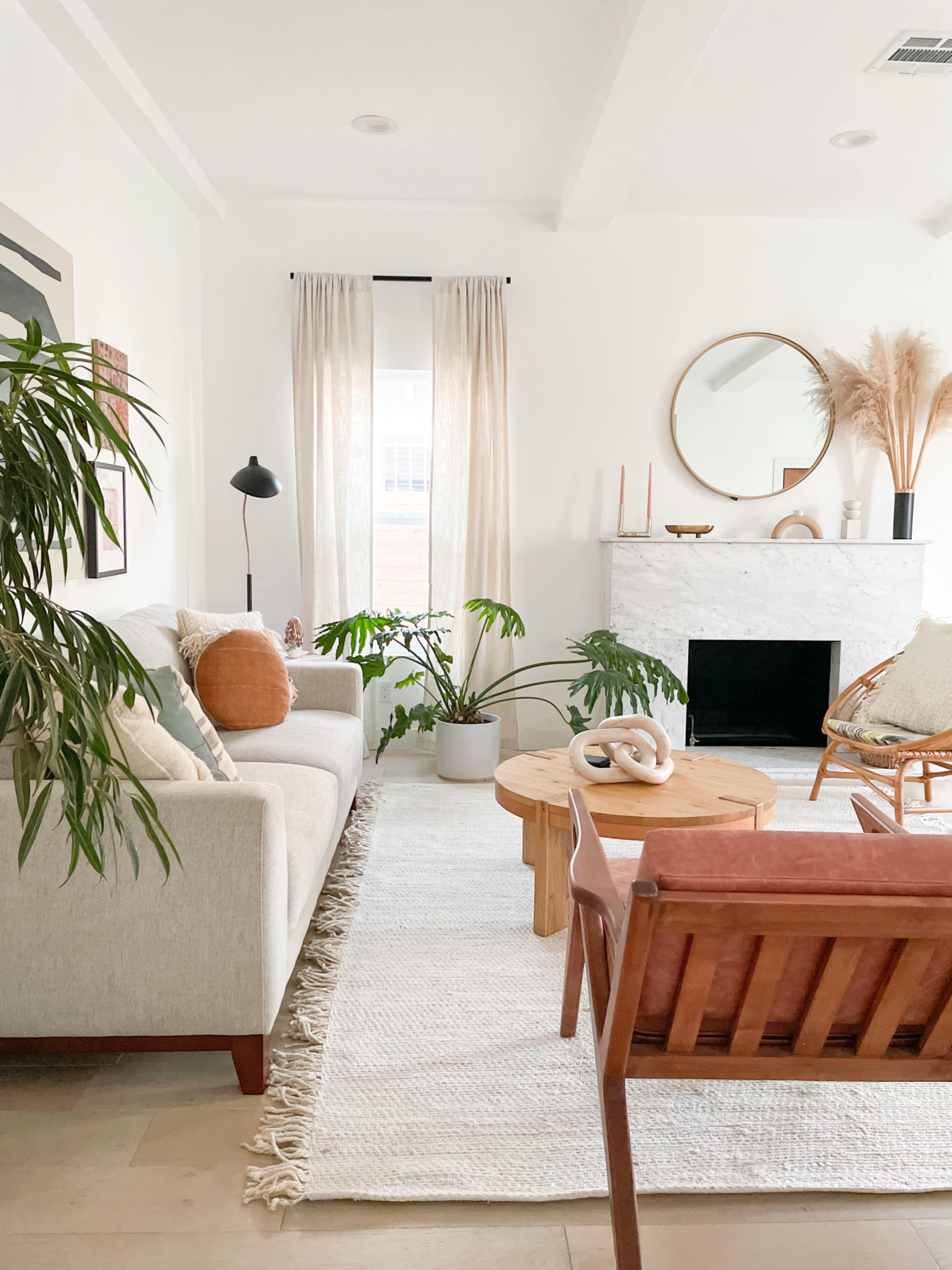 A bright living room features a light gray sofa, a wooden coffee table, and a decorative fireplace with plants and a round mirror above it.