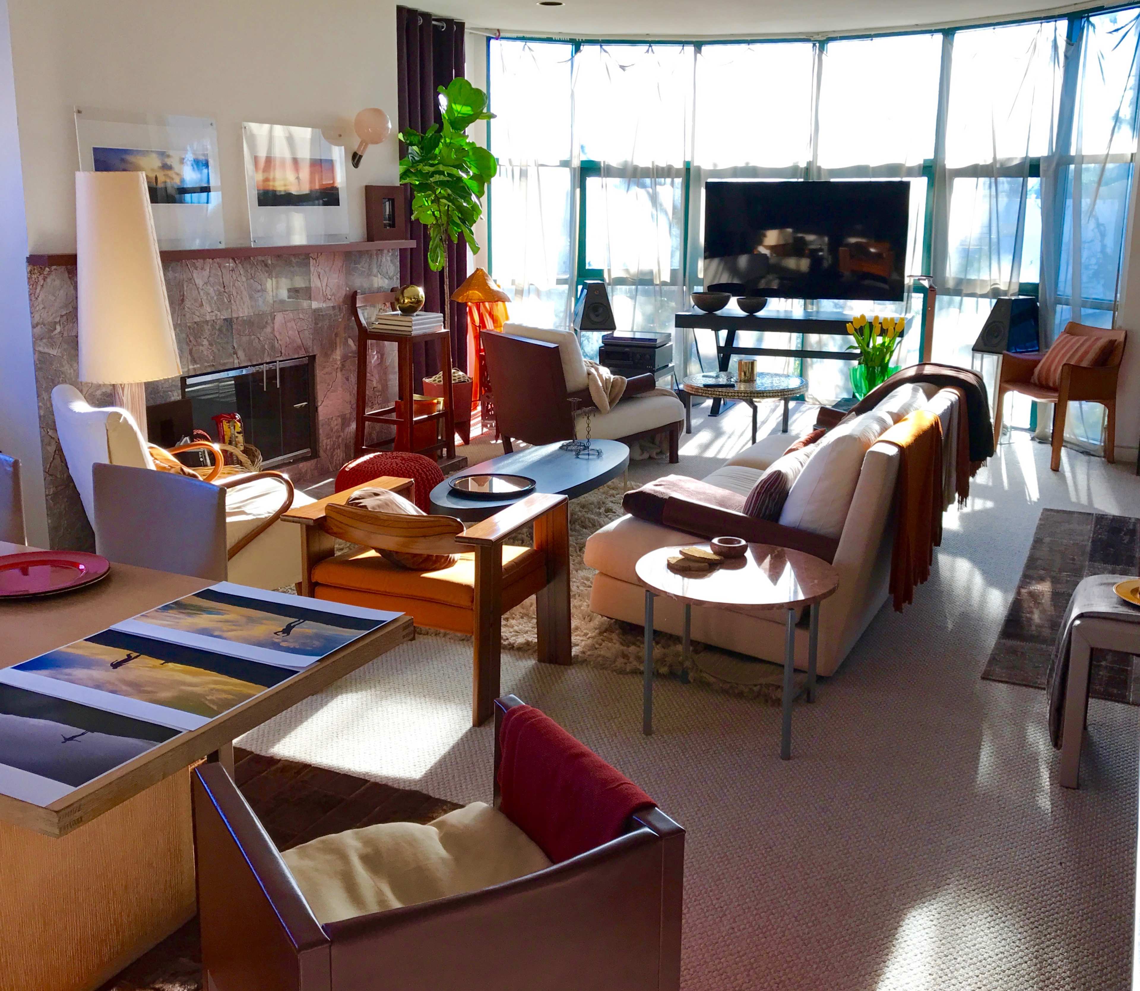 The image shows a modern living room with a fireplace, large windows, and various furniture pieces, including sofas, chairs, and a TV stand.