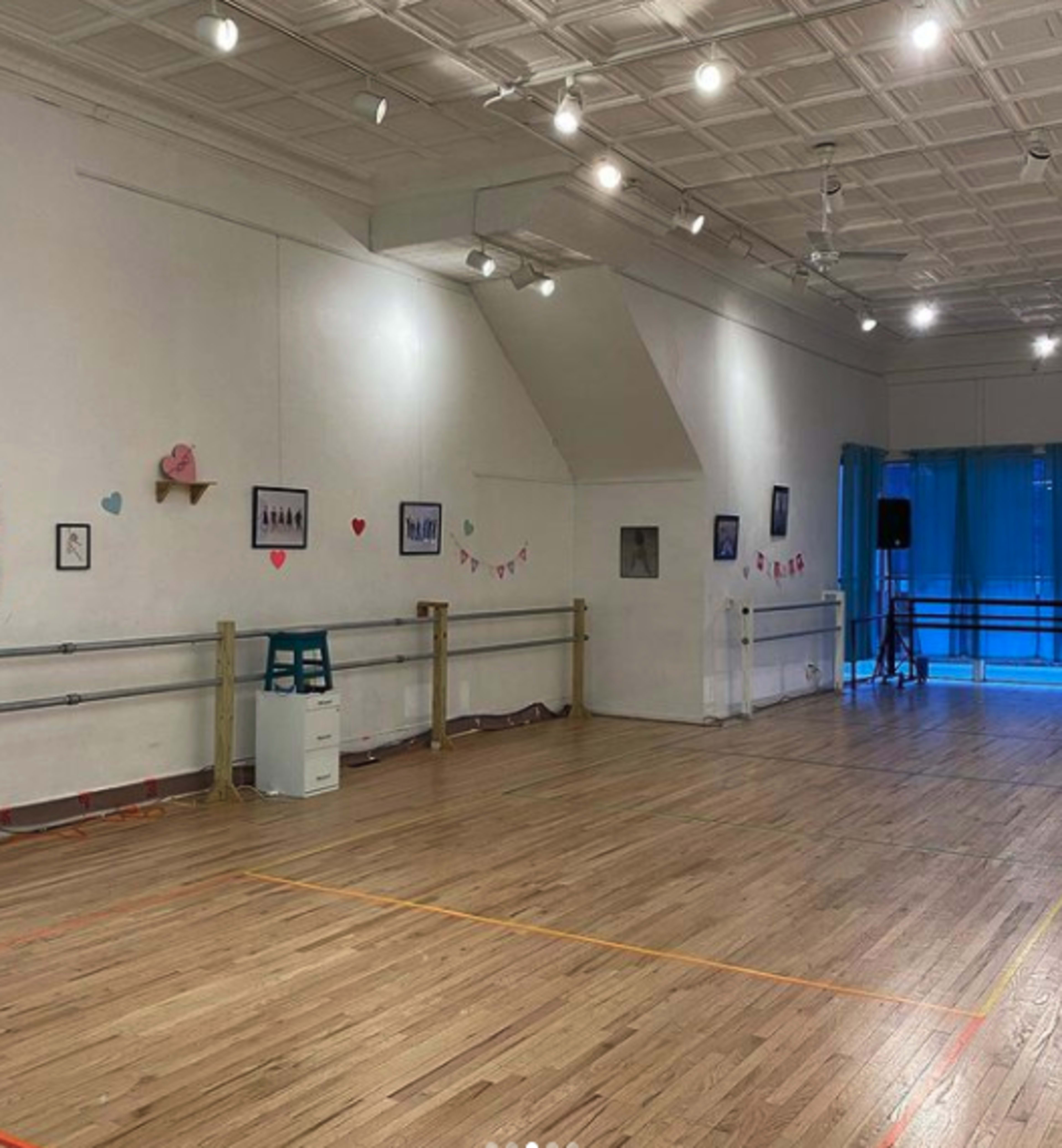 A spacious dance studio with wooden floors, decorated walls featuring framed pictures and heart-shaped decorations, and a large window with a blue curtain.