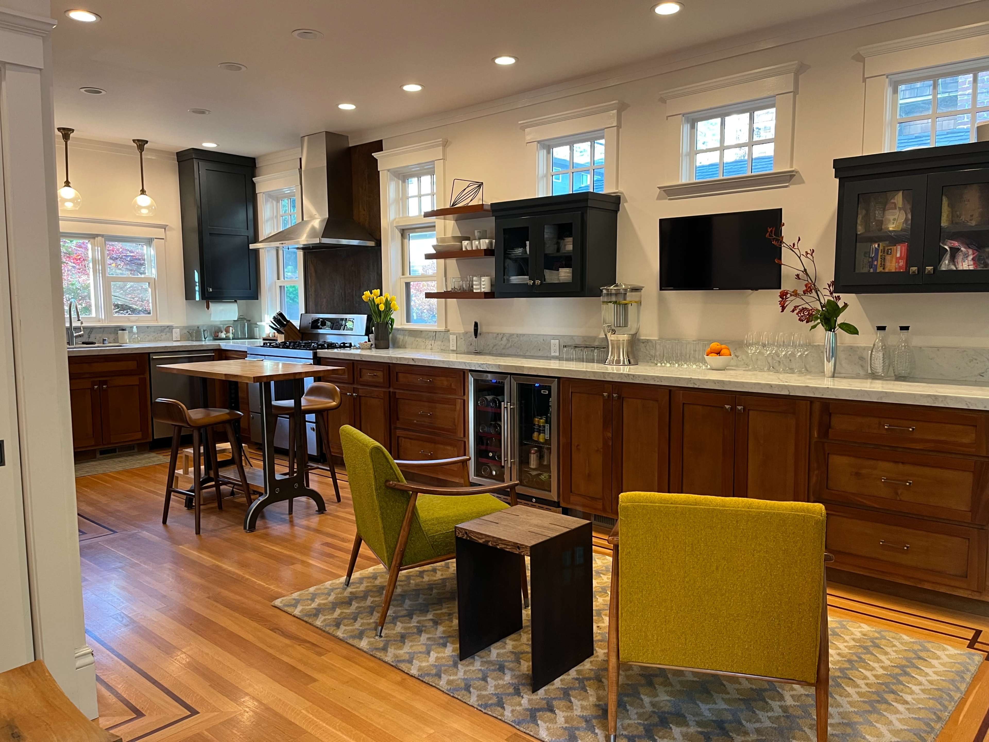 A modern kitchen with wooden cabinetry, a marble countertop, and a small seating area featuring two chairs and a side table.