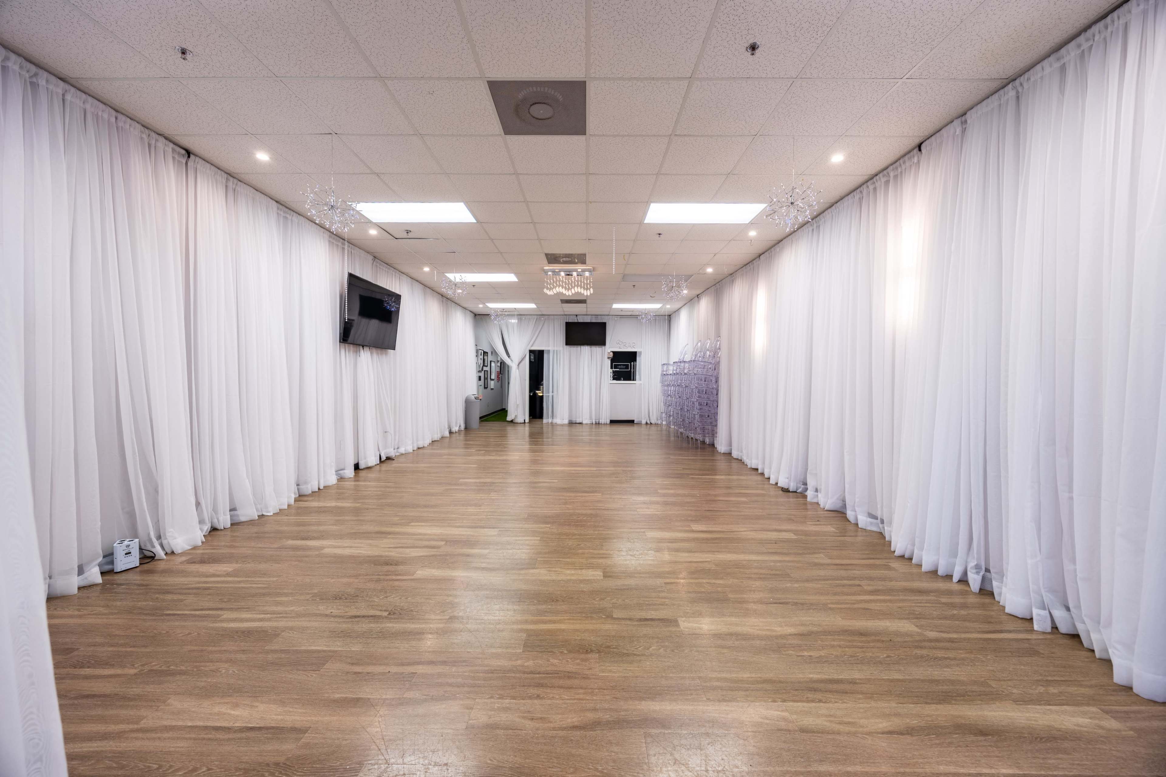The image shows a spacious, empty room with white drapes lining the walls and a wooden floor reflecting soft lighting.