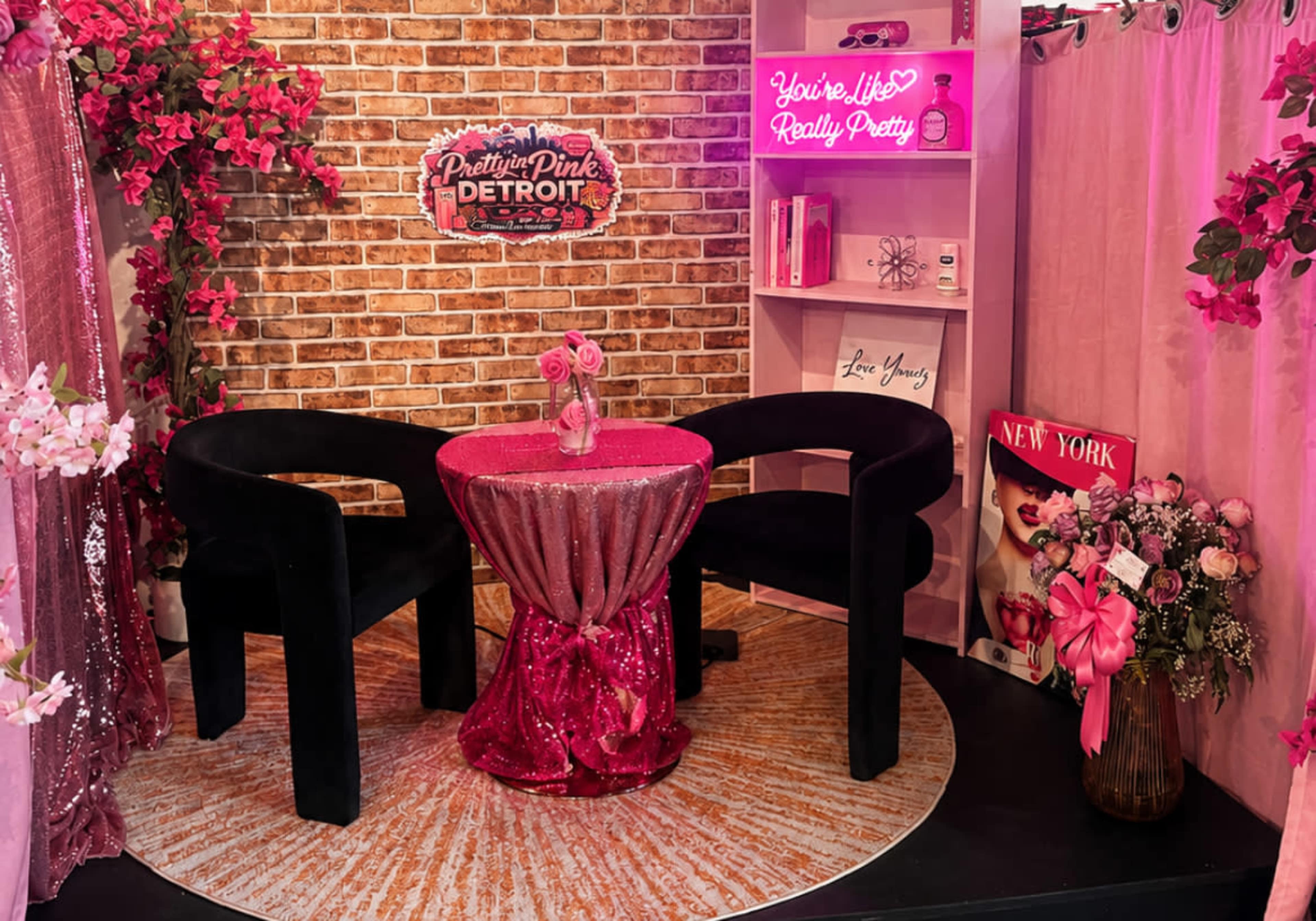 The image shows a vibrant indoor setting with a circular rug, a pink sequined tablecloth draped over a round table, two black chairs, and decorations featuring pink flowers and neon signs.