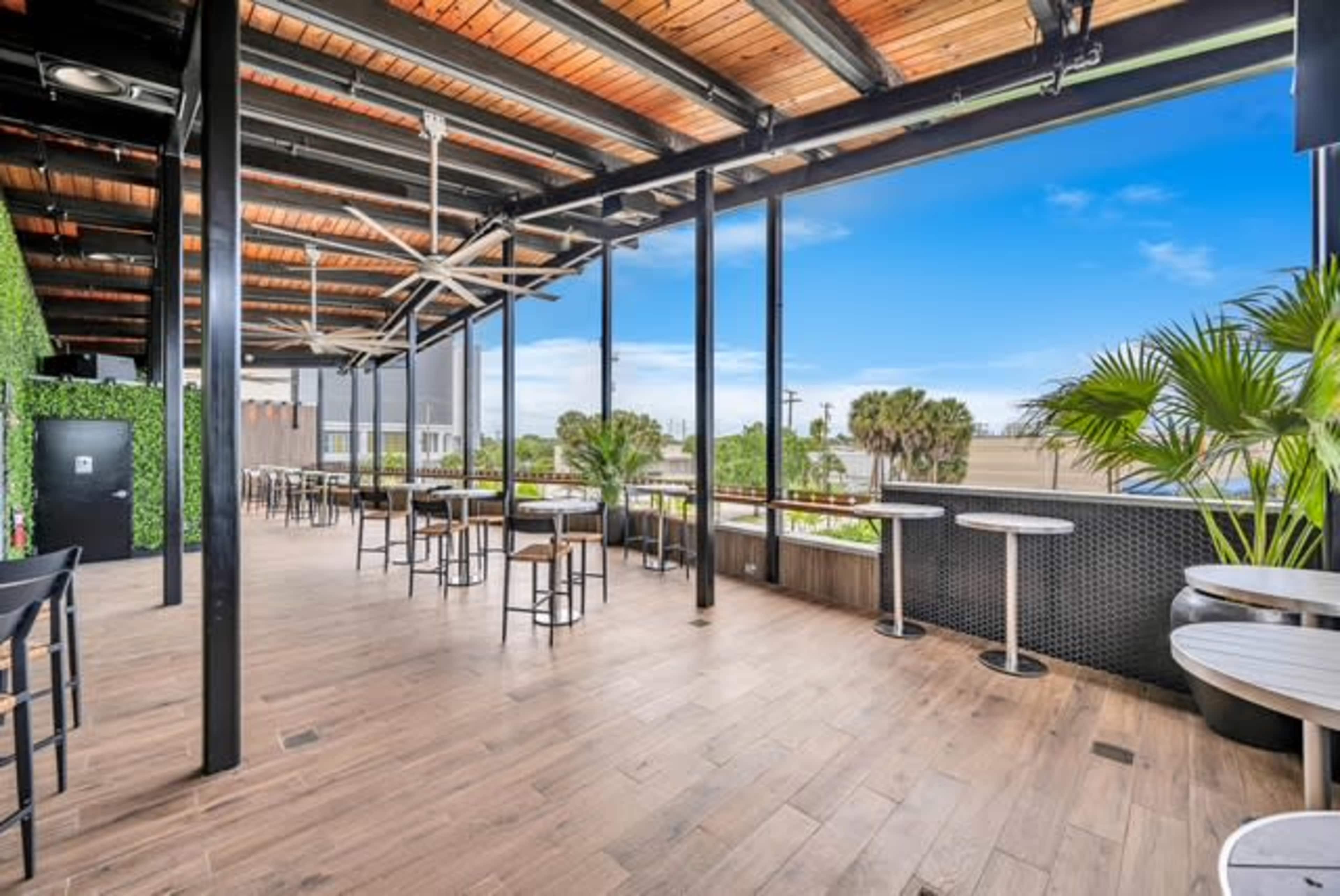 The image shows a spacious, modern bar area with wooden ceilings, large glass windows, and minimalistic furniture overlooking a landscaped outdoor setting.