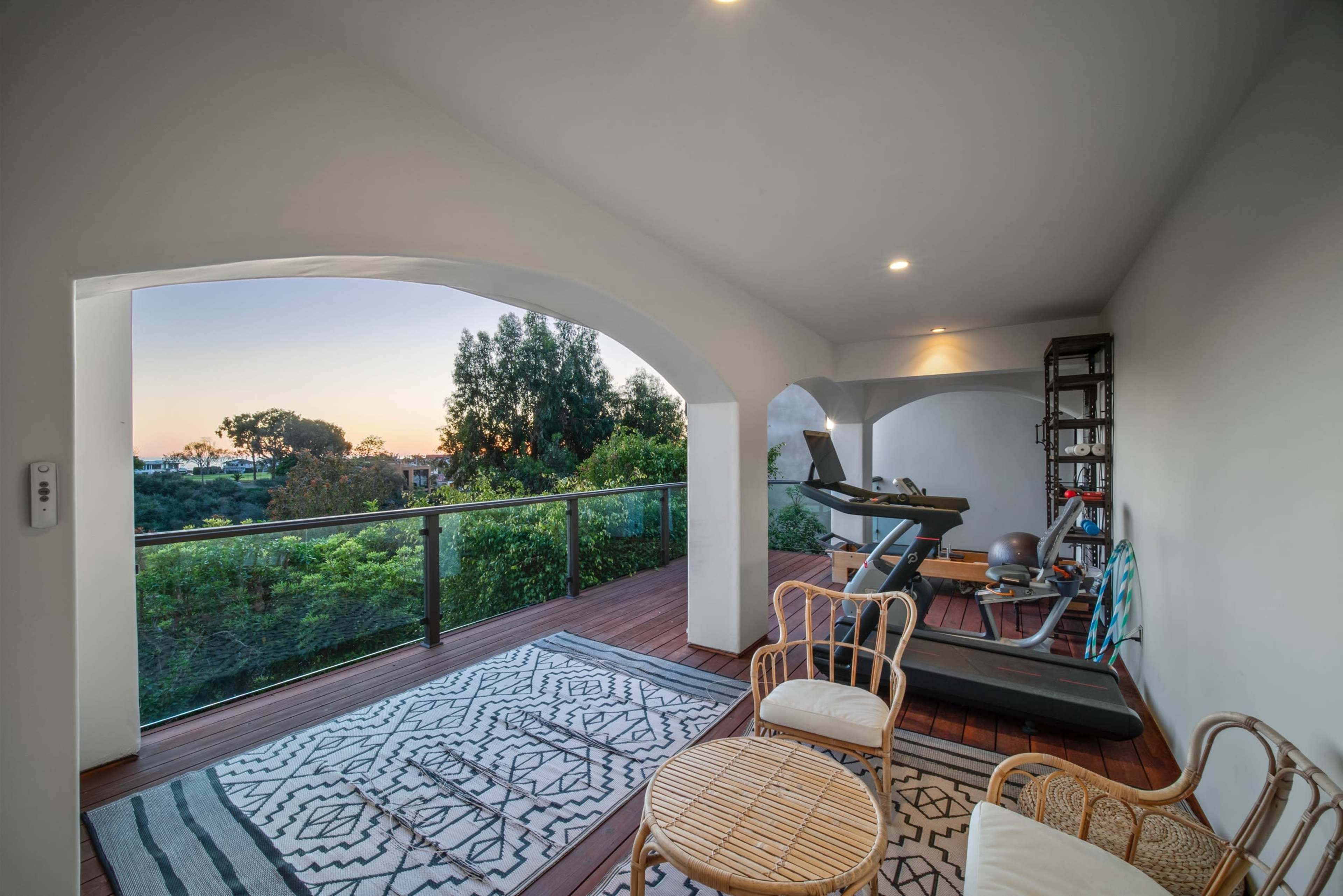 A modern balcony space furnished with a workout area, comfortable seating, and a view of greenery at sunset.