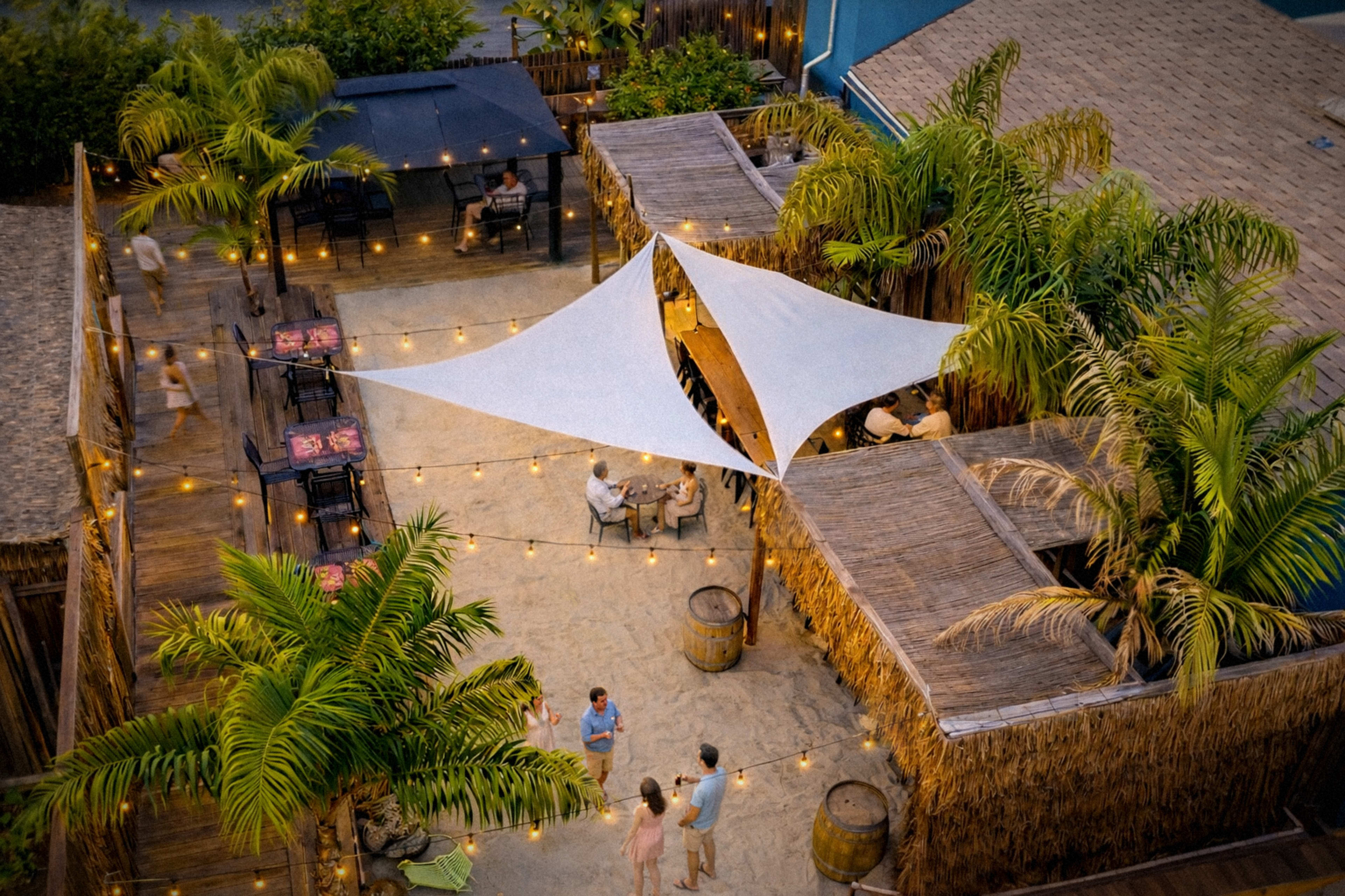 The image shows an outdoor area featuring several wooden structures, a white canopy, palm trees, and string lights, with people gathered socializing.
