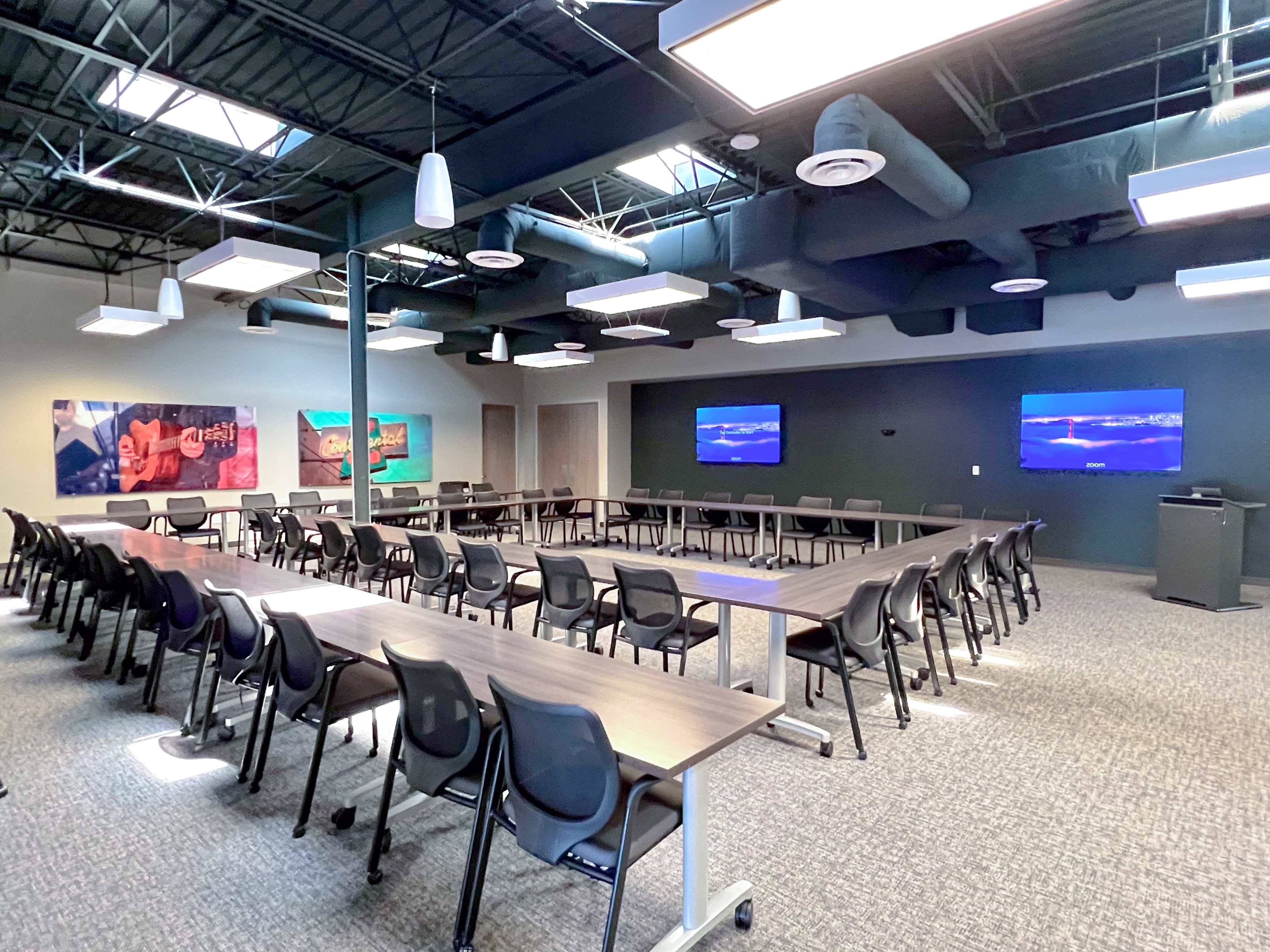 A modern conference room features several rows of chairs organized around tables, with wall-mounted screens and artwork displayed on the walls.