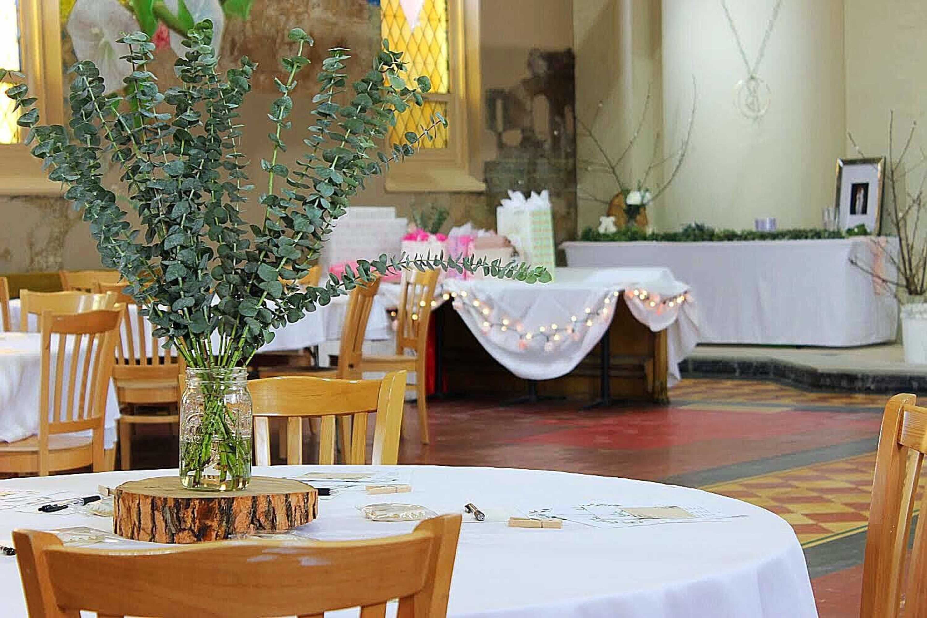 A dining area features round tables with white tablecloths, each adorned with a vase of eucalyptus and a wooden slice, while a gift table is set up in the background.