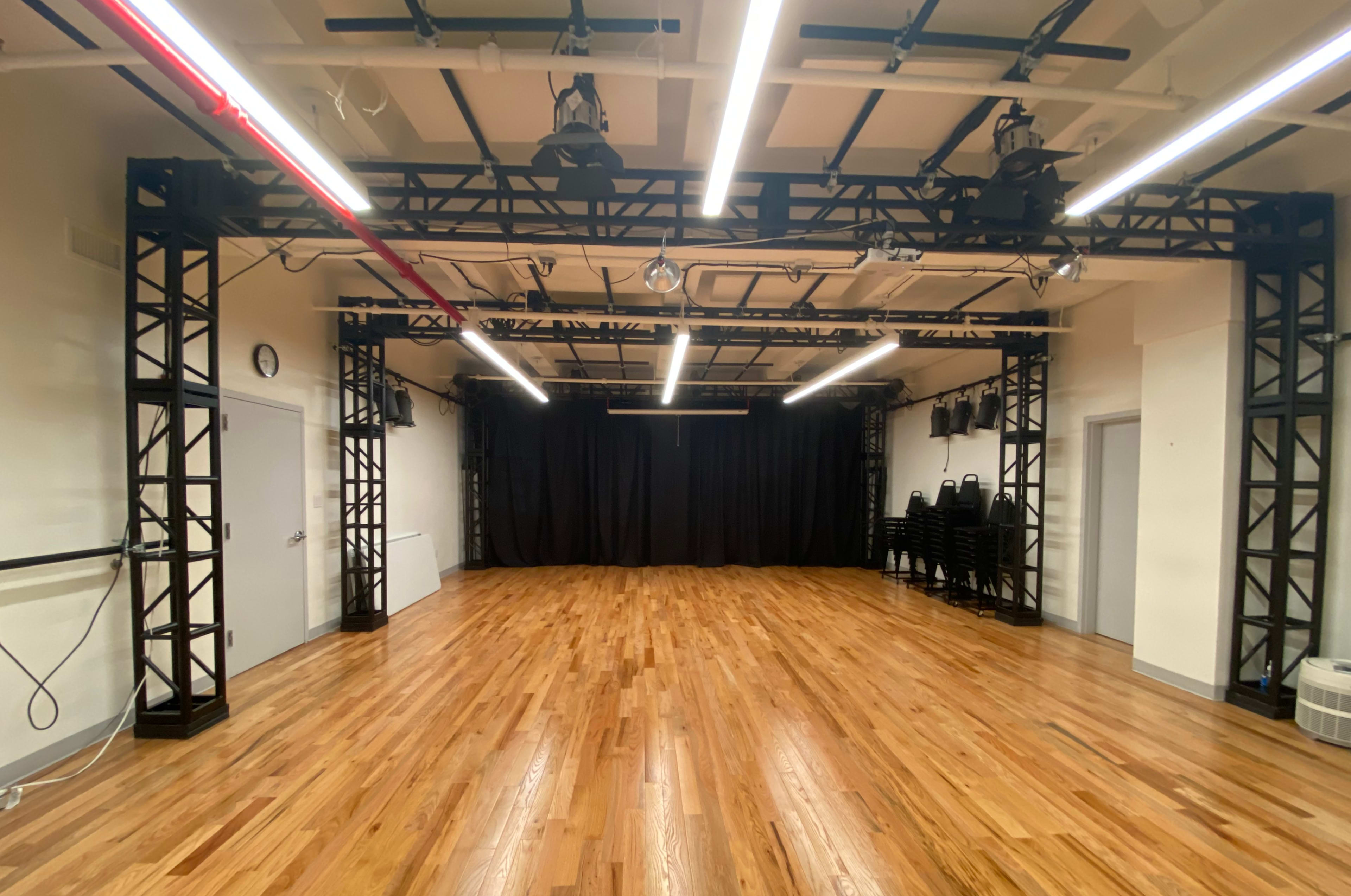 A spacious wooden-floored rehearsal studio with black support beams, overhead lights, and black curtains at one end.