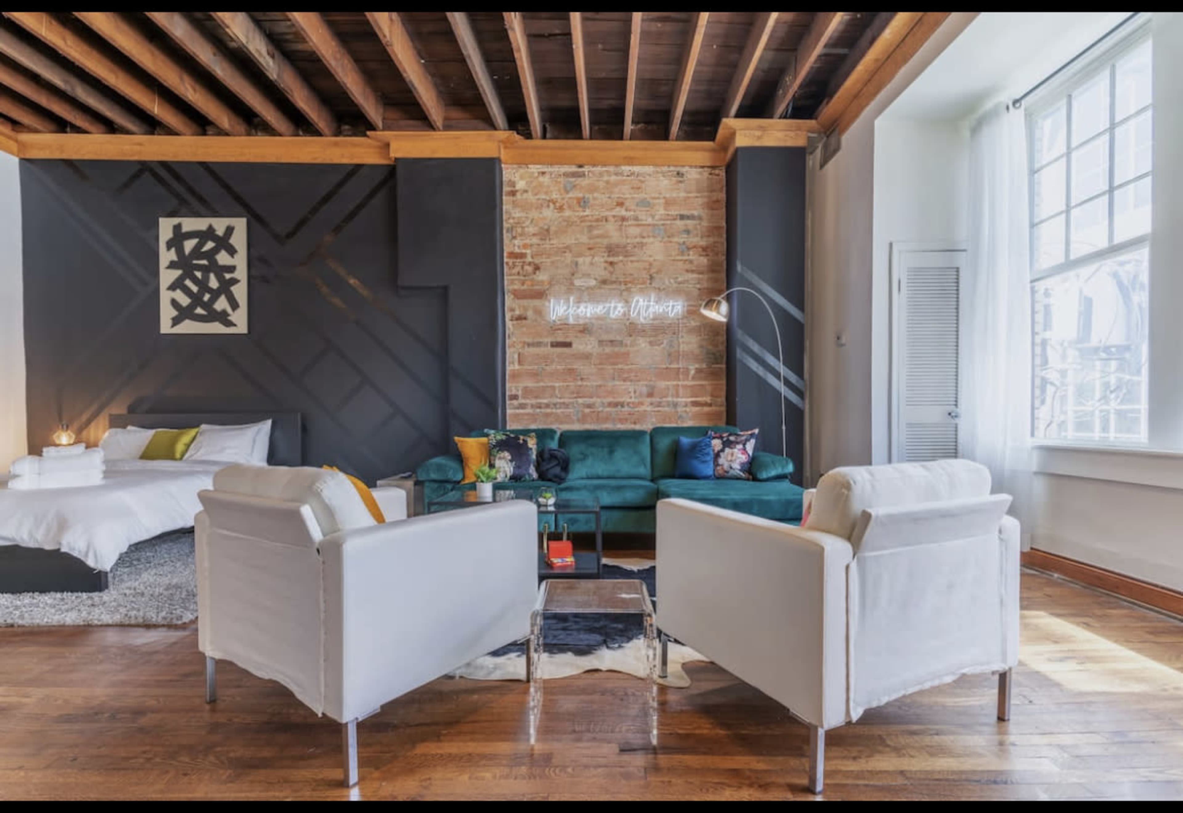 A stylish living space with a green sofa, two white chairs, a coffee table, and a bed visible in the background, set against a brick wall and wooden ceiling.