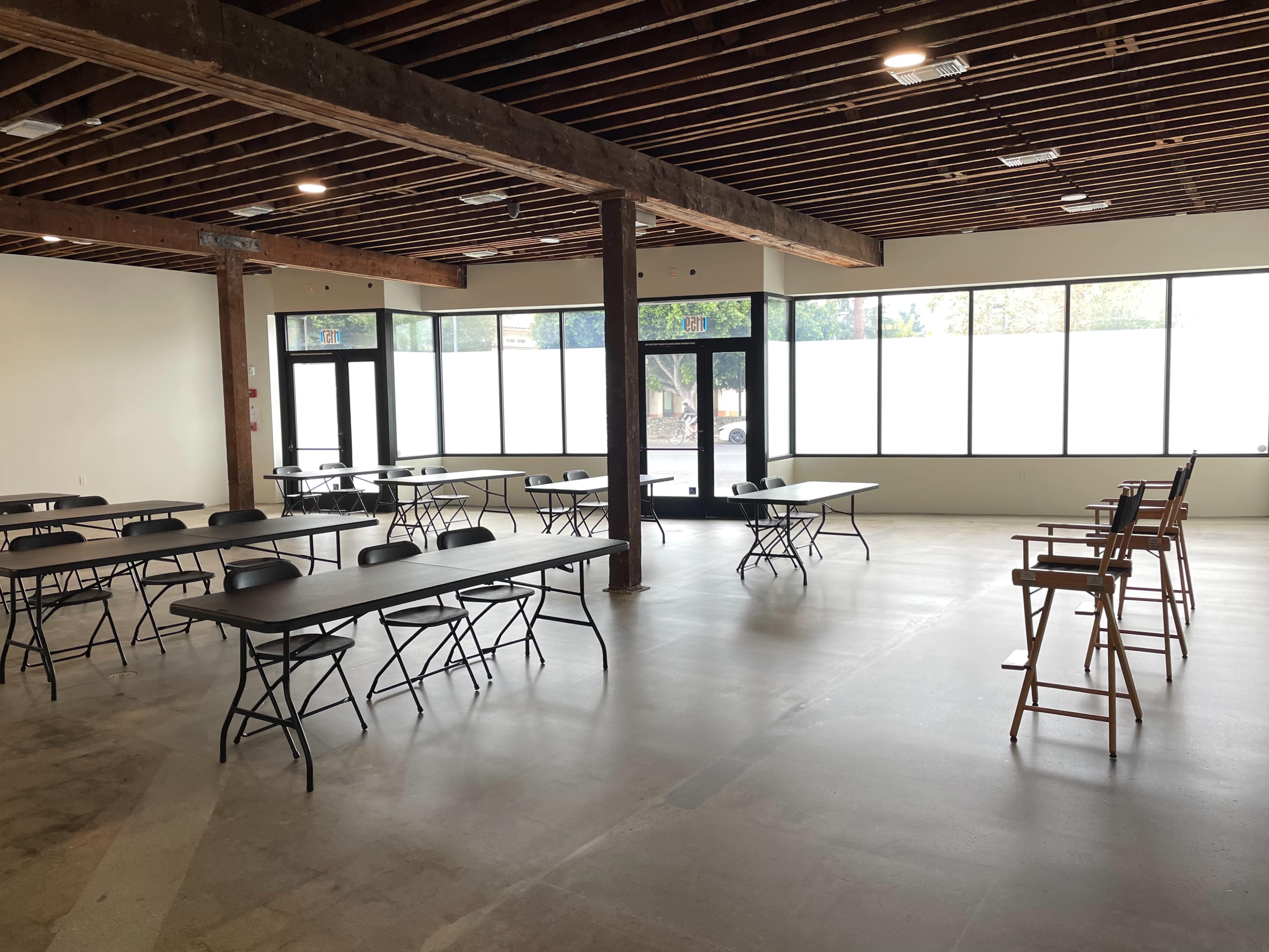 An empty room with several black folding tables and chairs arranged in a spacious layout under wooden beams and large windows.