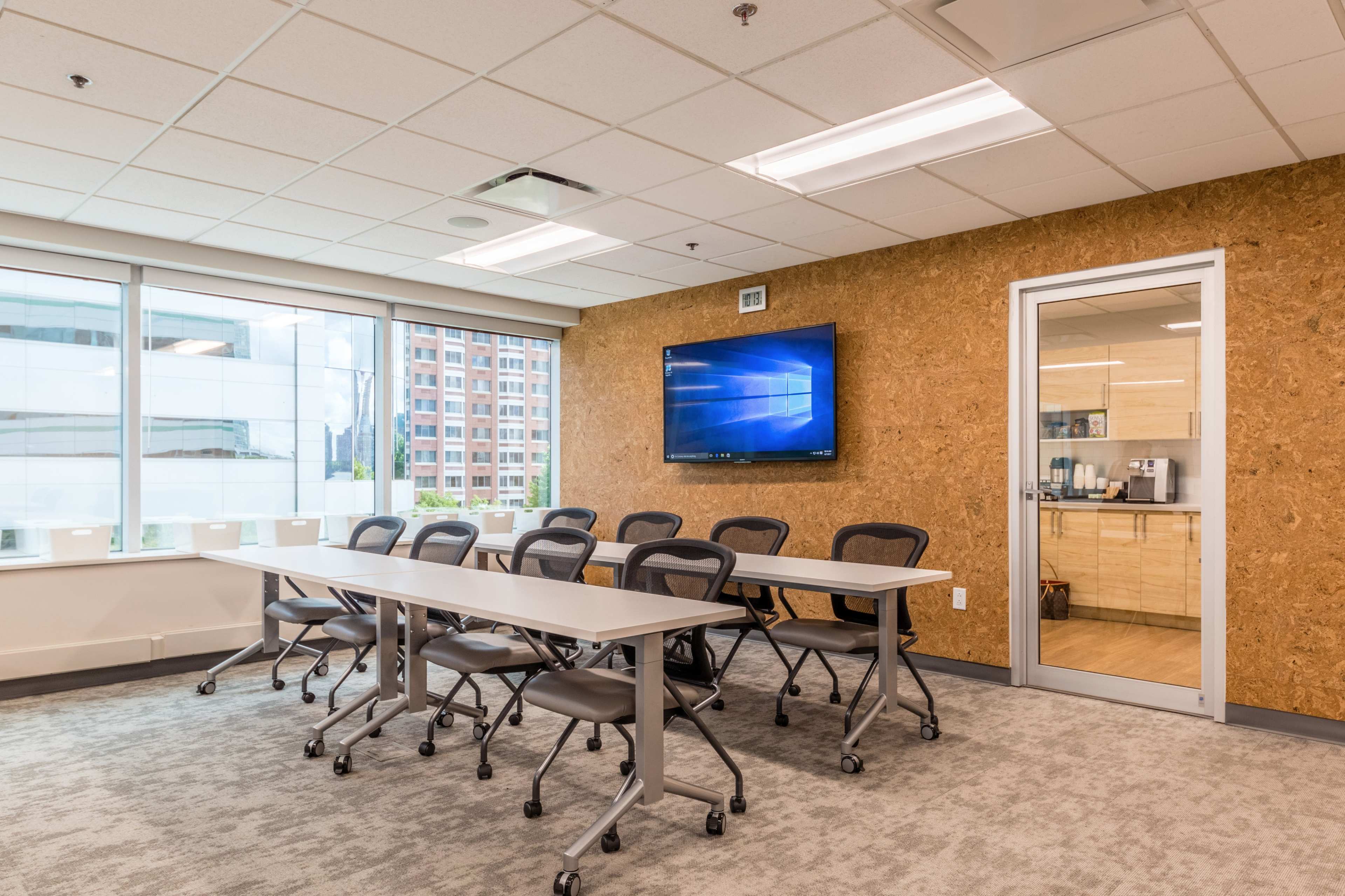 A modern conference room features a long table with rolling chairs, a large screen mounted on the wall, and windows offering a view of a cityscape.