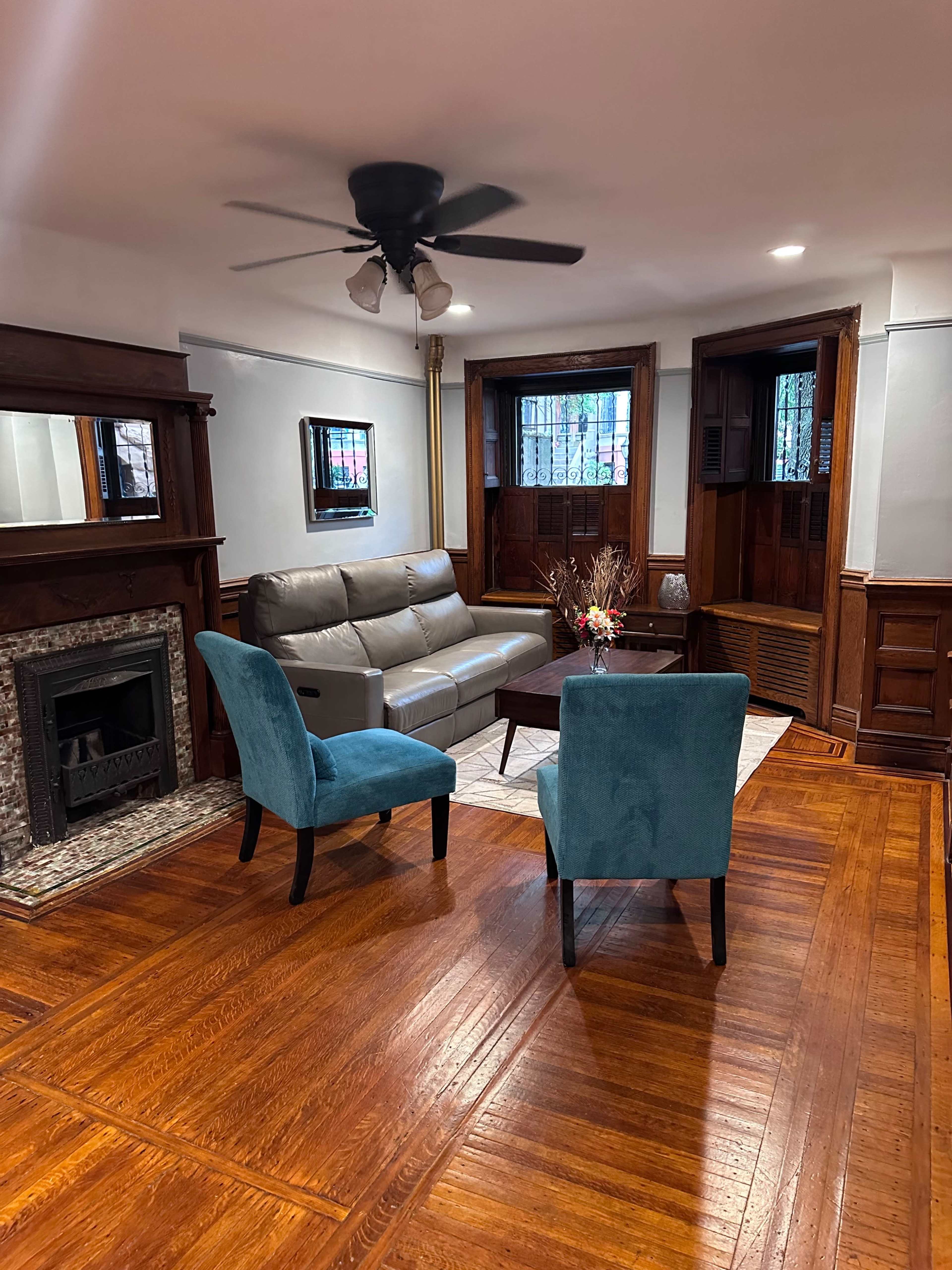 The image shows a living room with a gray sofa, two teal chairs, a small round table, and a decorative fireplace, all set on a polished wooden floor.