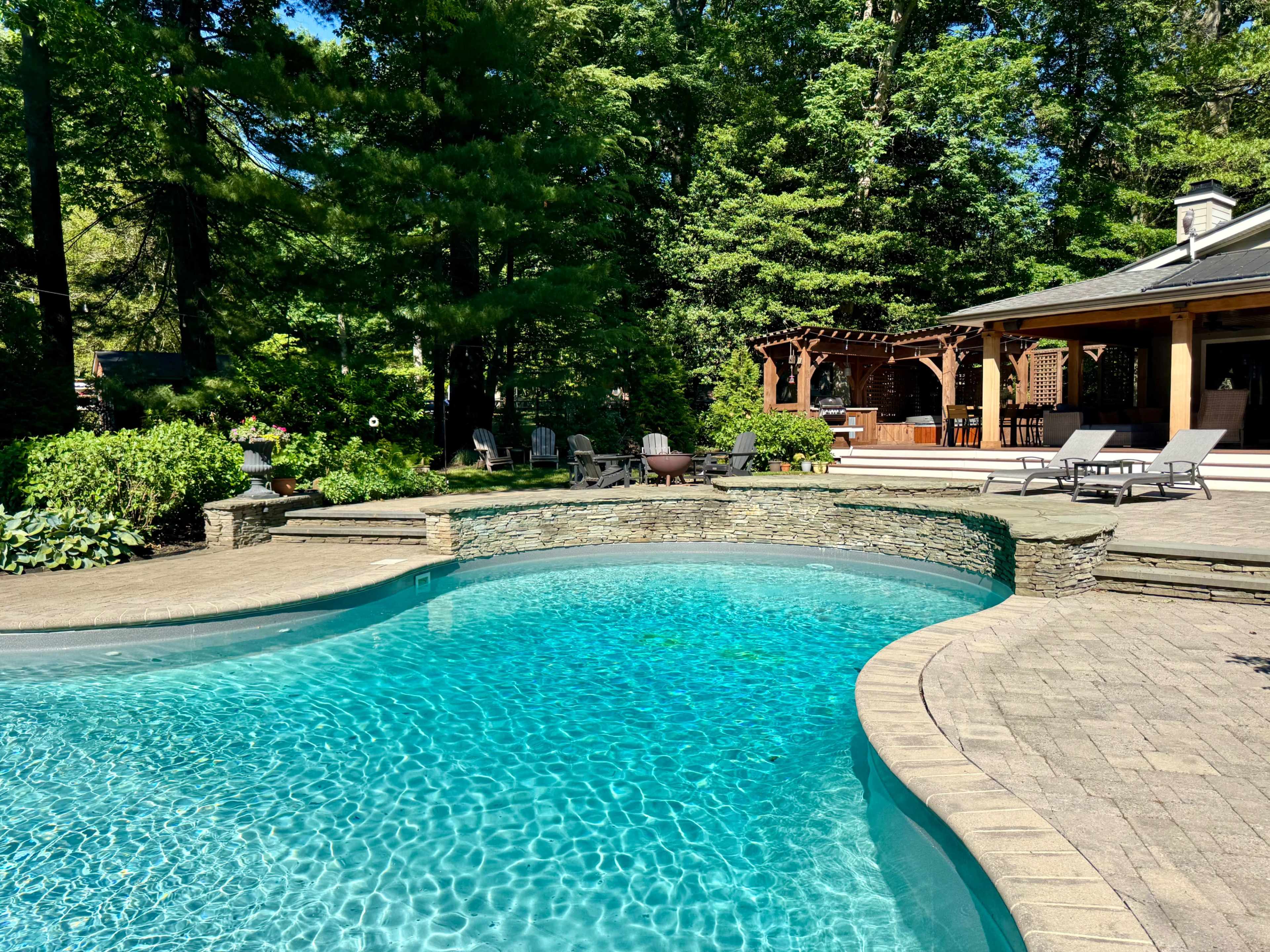 The image shows a landscaped backyard with a clear swimming pool surrounded by various seating areas and a covered patio.