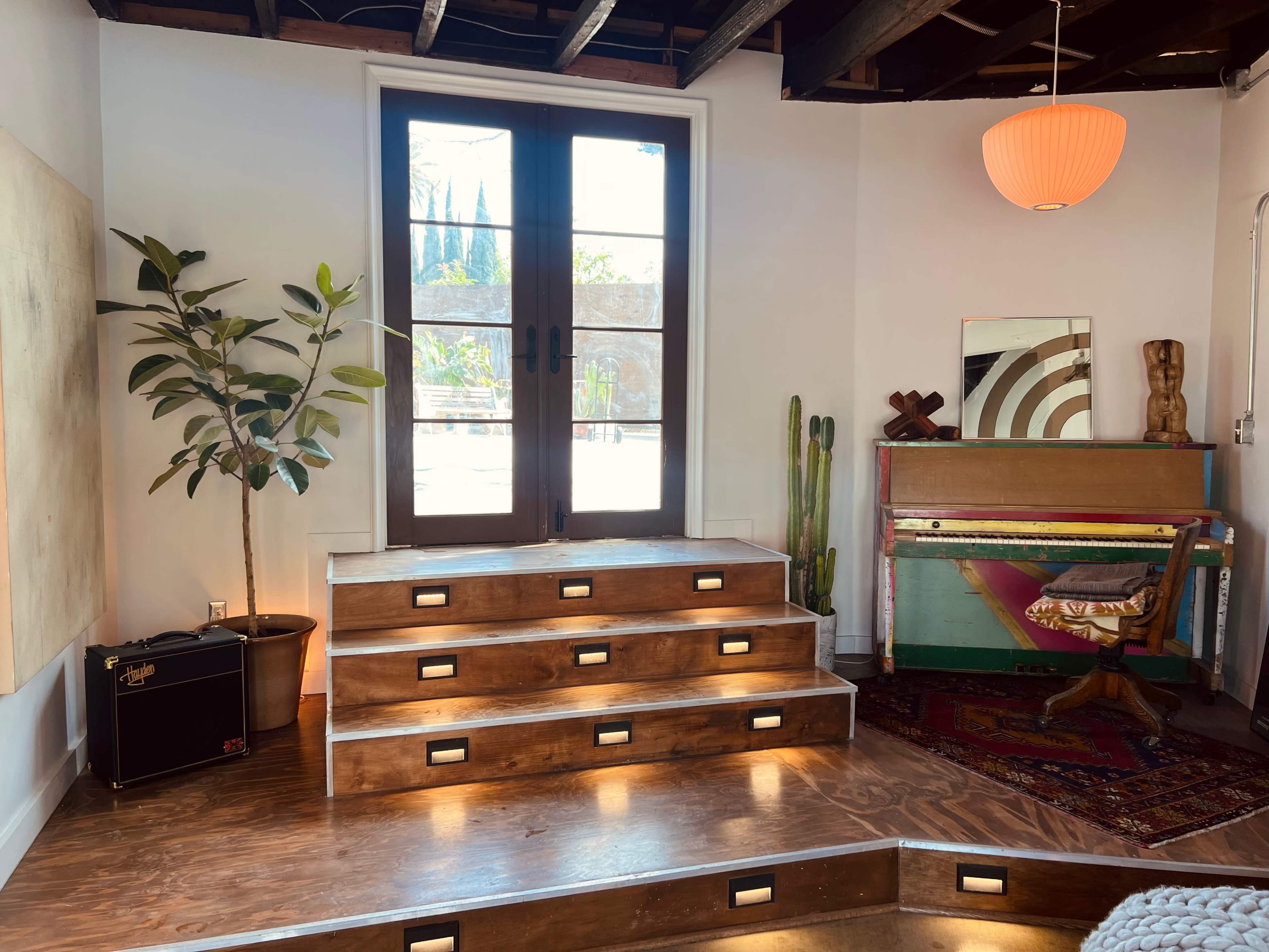 The scene features a room with wooden steps leading to glass double doors, a potted plant, a colorful vintage piano, and a wooden sculpture against the walls.