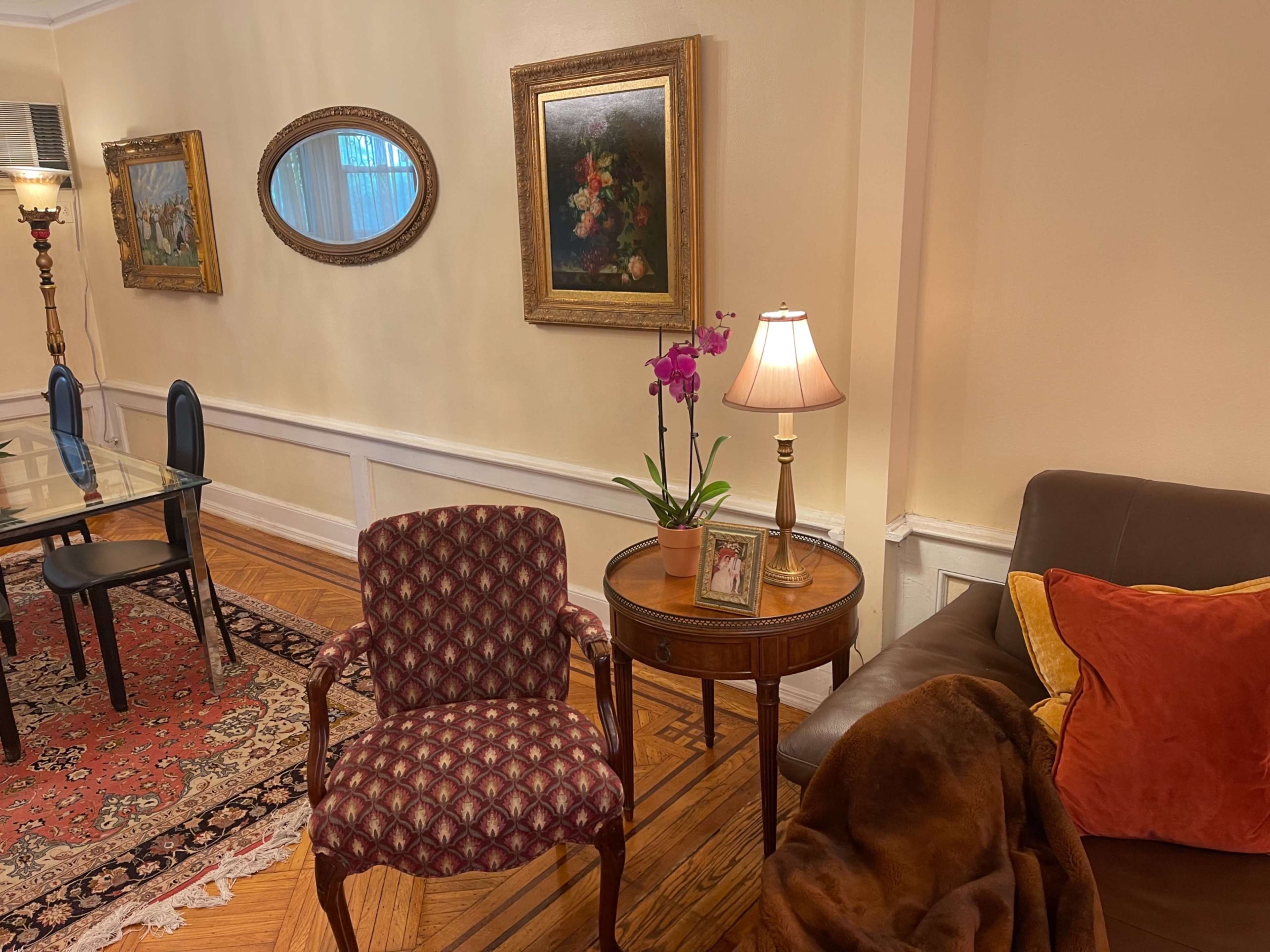 A cozy living room with a patterned chair, a side table featuring a lamp and an orchid, and framed artwork on the walls.