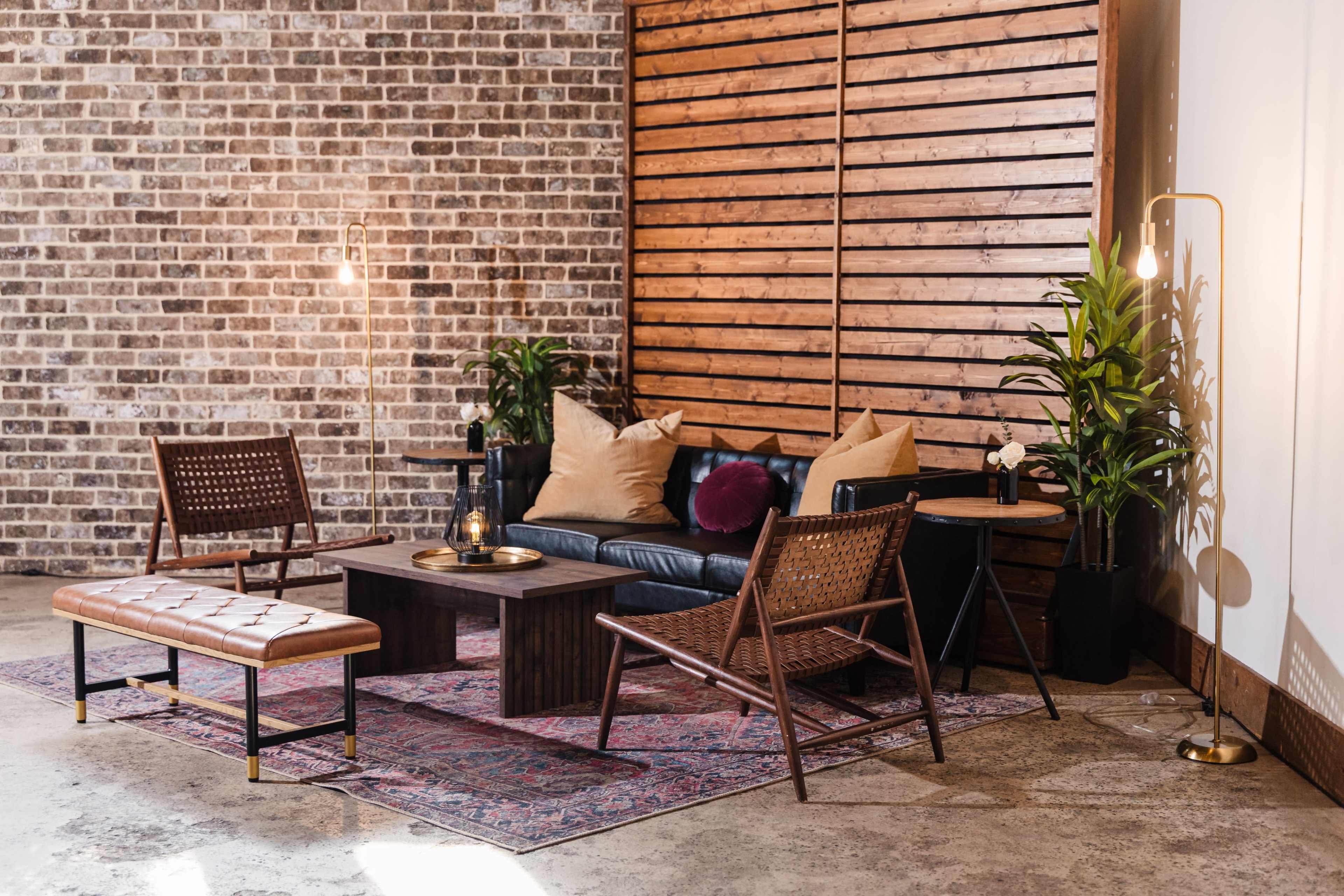 The image shows a modern living room setup featuring a black leather sofa, two woven chairs, a wooden coffee table, and decorative plants against a textured brick wall.