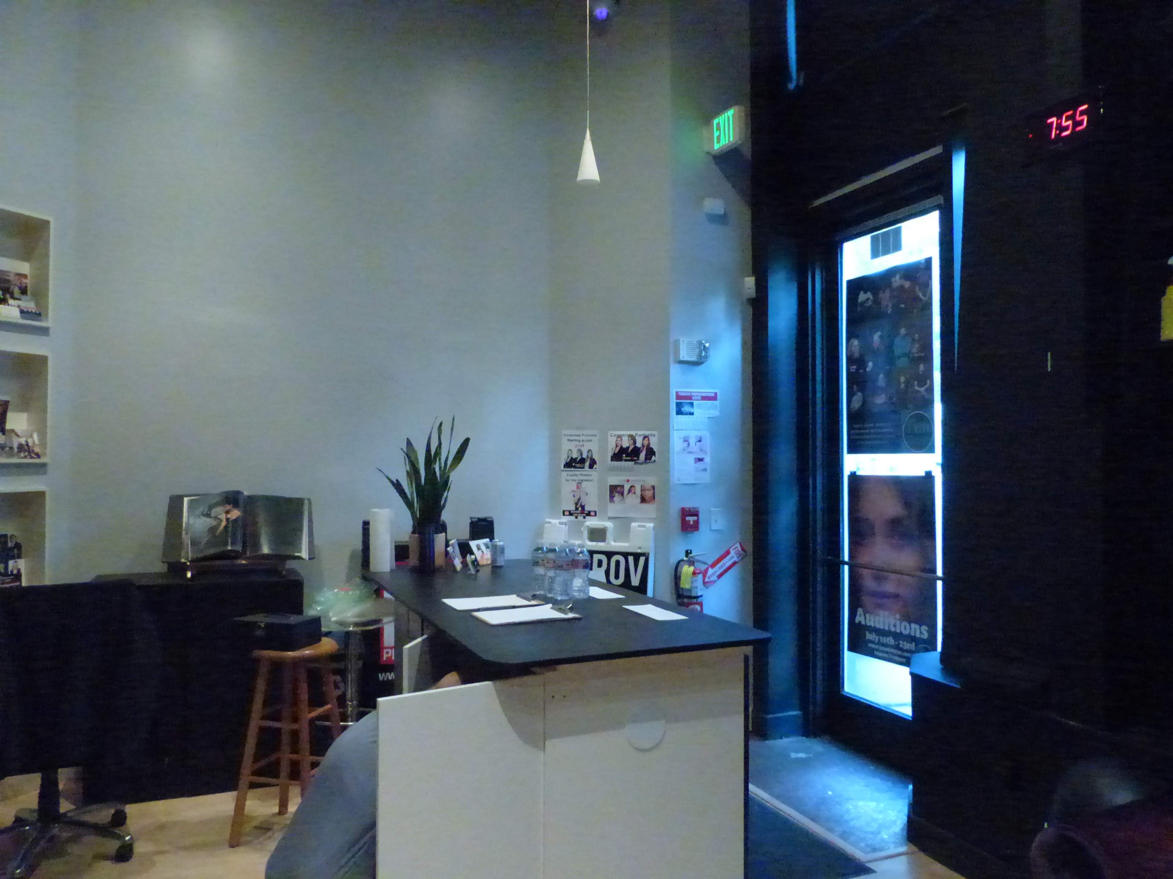 A dimly lit room with a small bar area, a stool, a potted plant, and an open door revealing posters and a clock displaying the time as 7:55.
