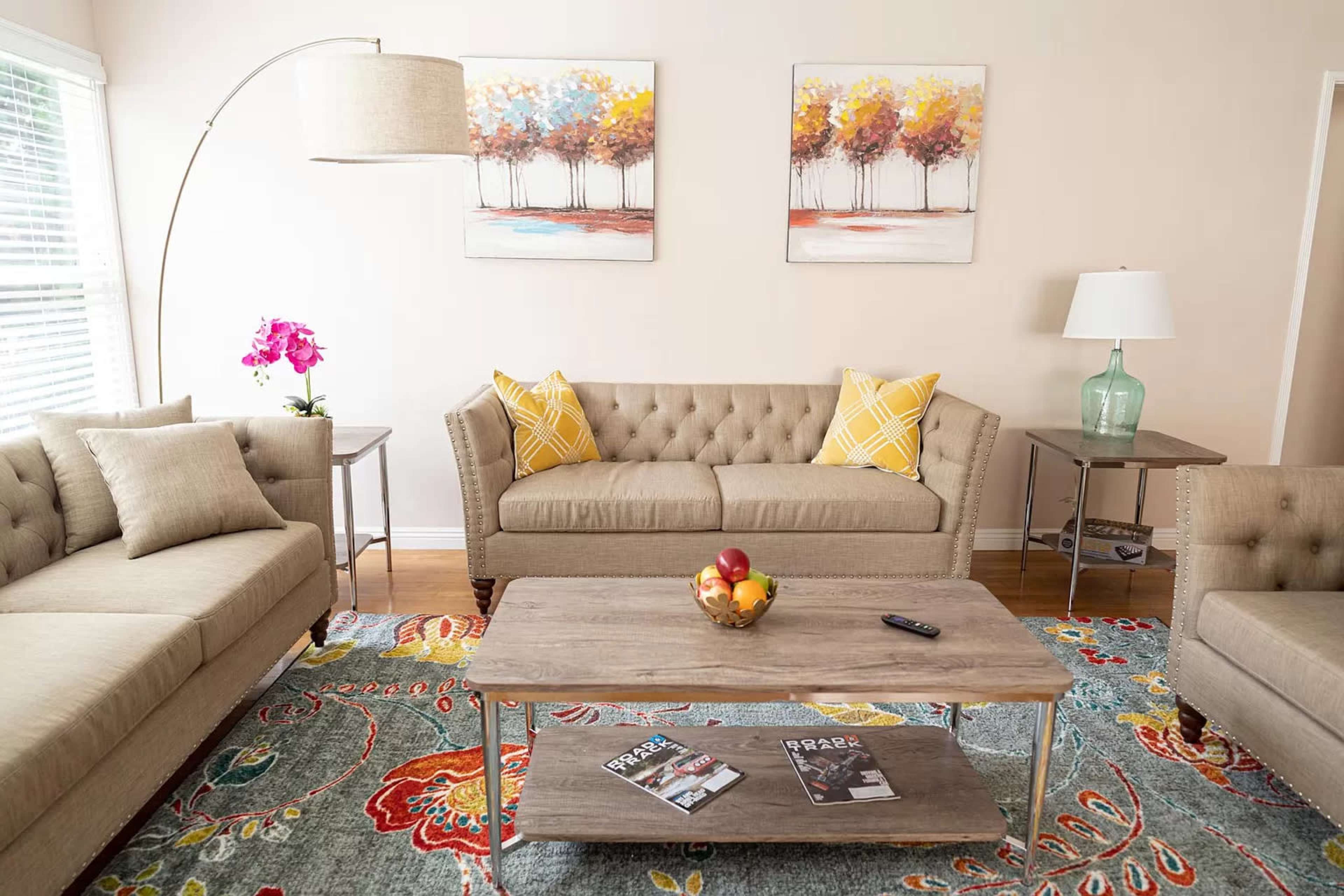 The image shows a cozy living room with a light-colored sofa set, a wooden coffee table with magazines, and decorative wall art, all arranged on a colorful area rug.