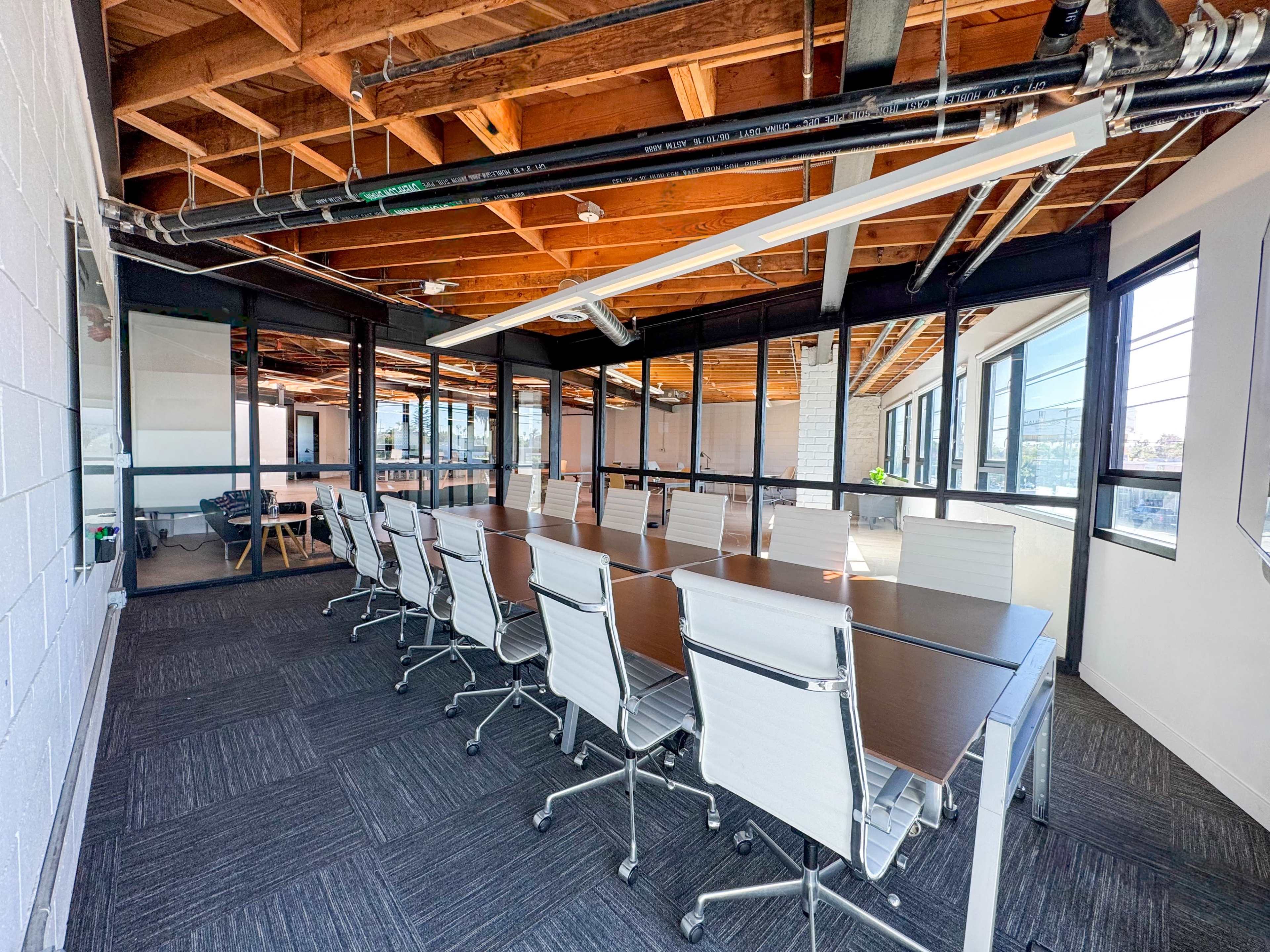 A spacious conference room features a long table surrounded by white rolling chairs, with large windows and exposed wooden beams overhead.