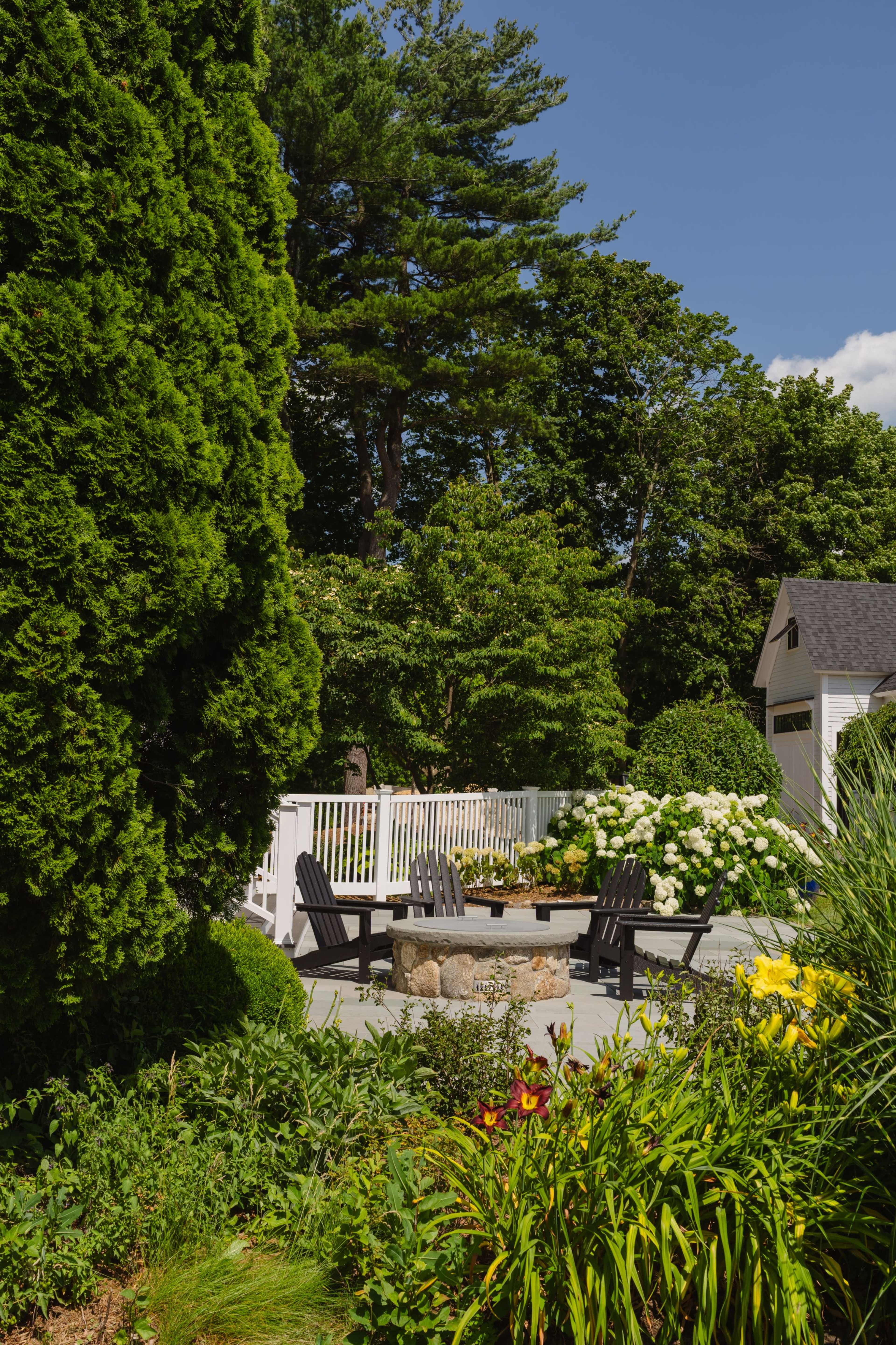 A circular stone fire pit is surrounded by black adirondack chairs in a backyard with green foliage and a white fence.