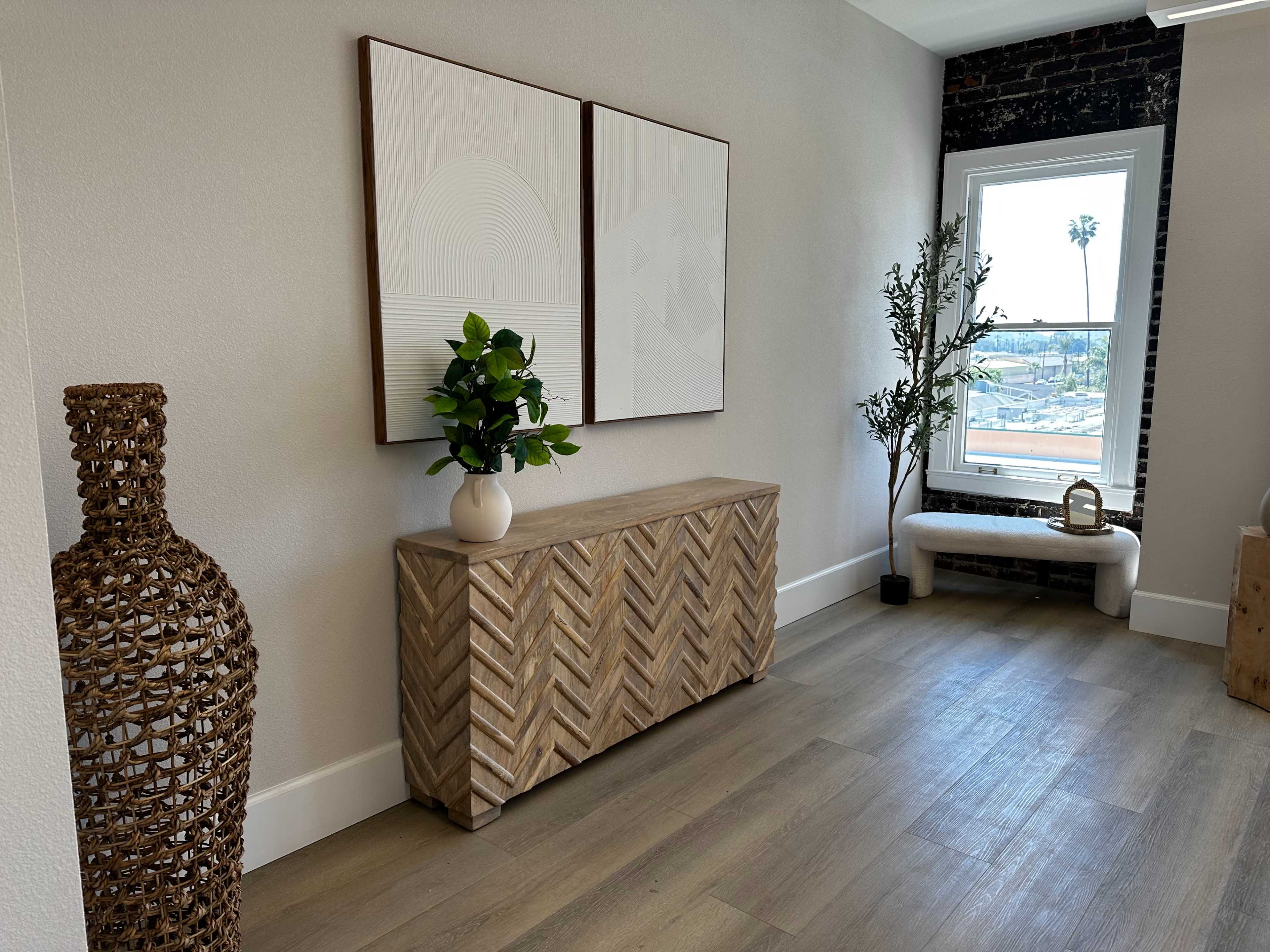 A modern interior space featuring a wooden cabinet with a herringbone pattern, two framed artworks on the wall, and a window with a view of palm trees outside.
