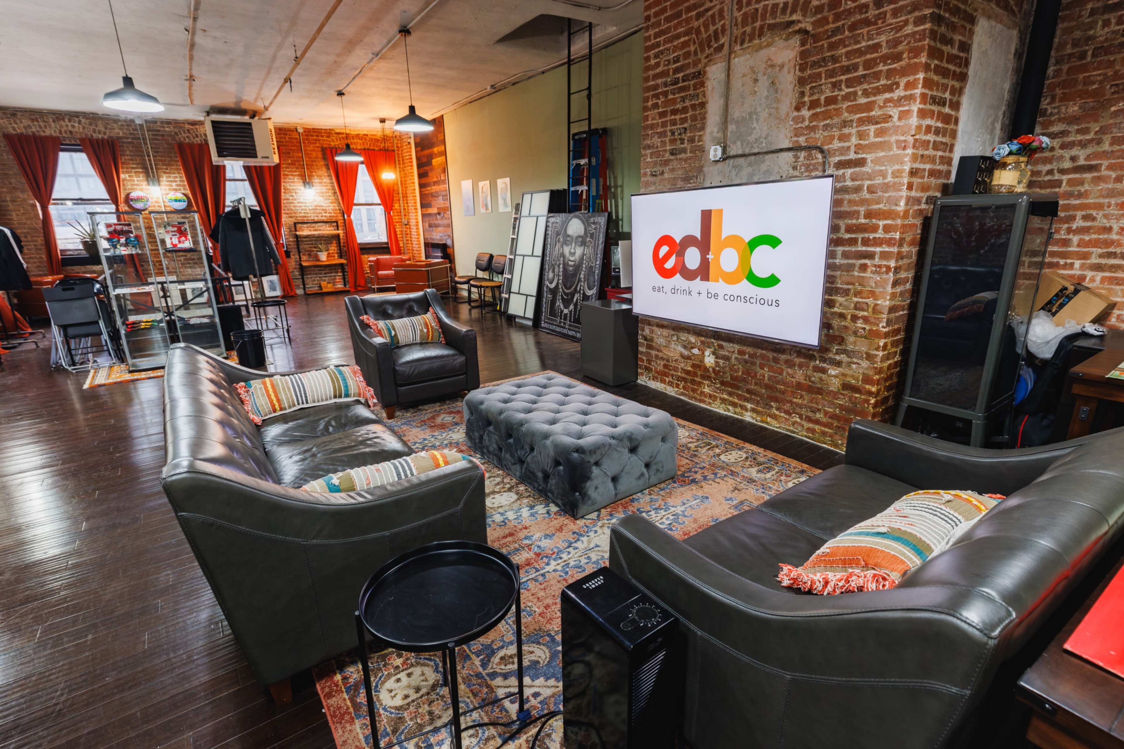 The image shows a modern, spacious lounge area with brick walls, featuring leather sofas, a tufted ottoman, and a large screen displaying the logo for "edbc" against a backdrop of colorful artwork and decor.