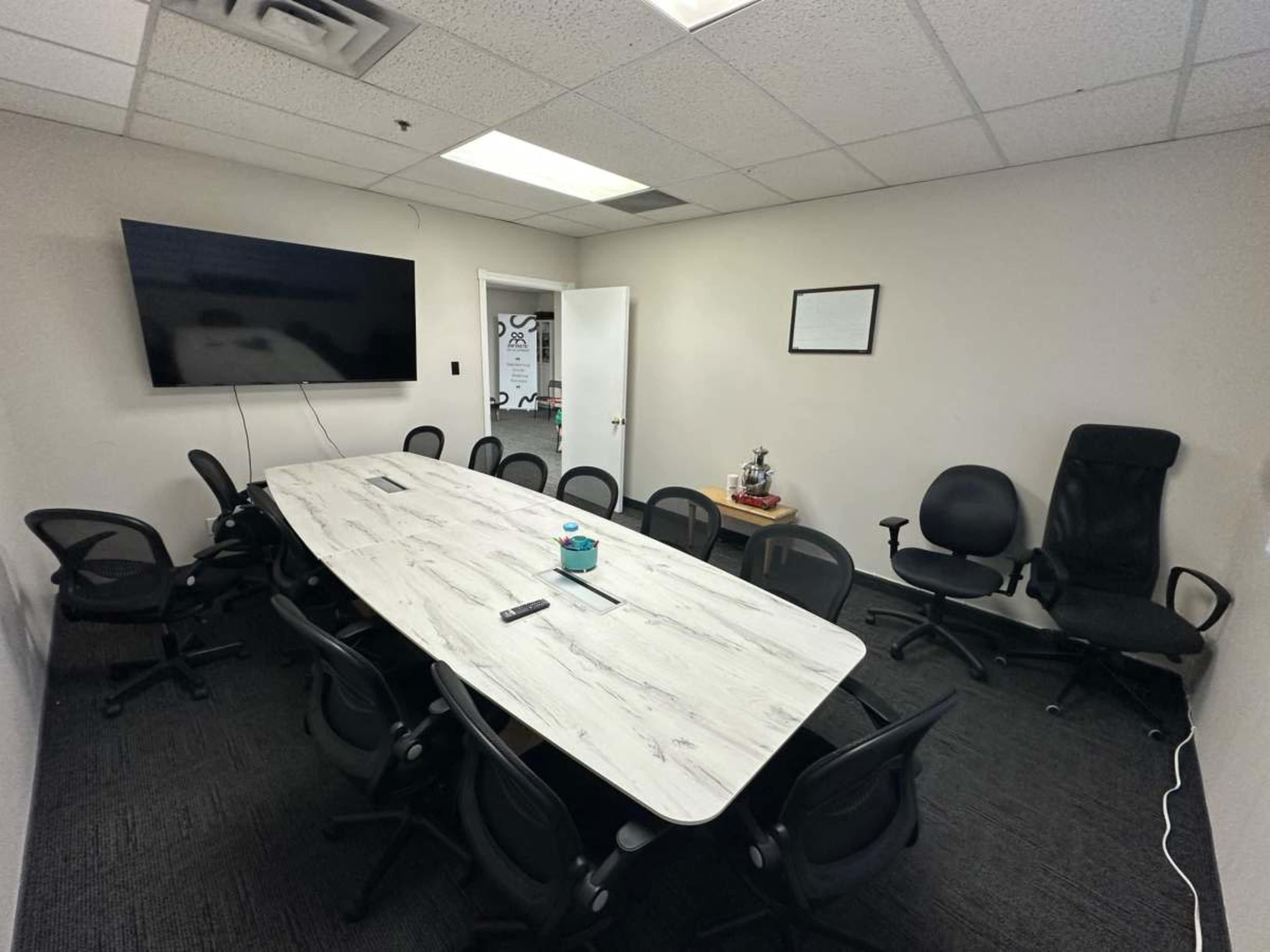 The image shows a conference room with a long, rectangular table surrounded by several office chairs, a television mounted on the wall, and a door leading to another room.