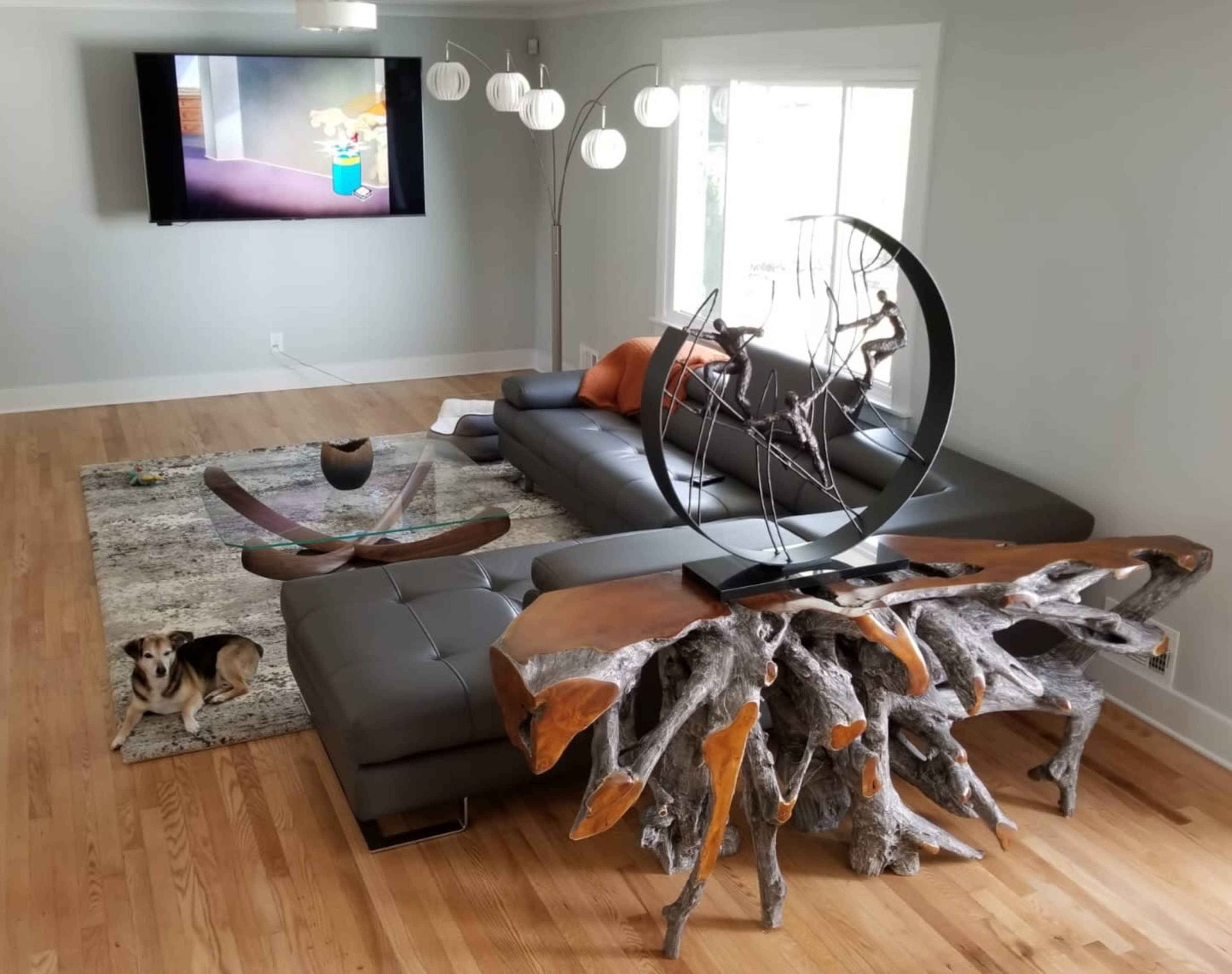 A modern living room with a gray sectional sofa, a unique wooden coffee table, a decorative sculpture, a beige area rug, and a television mounted on the wall.