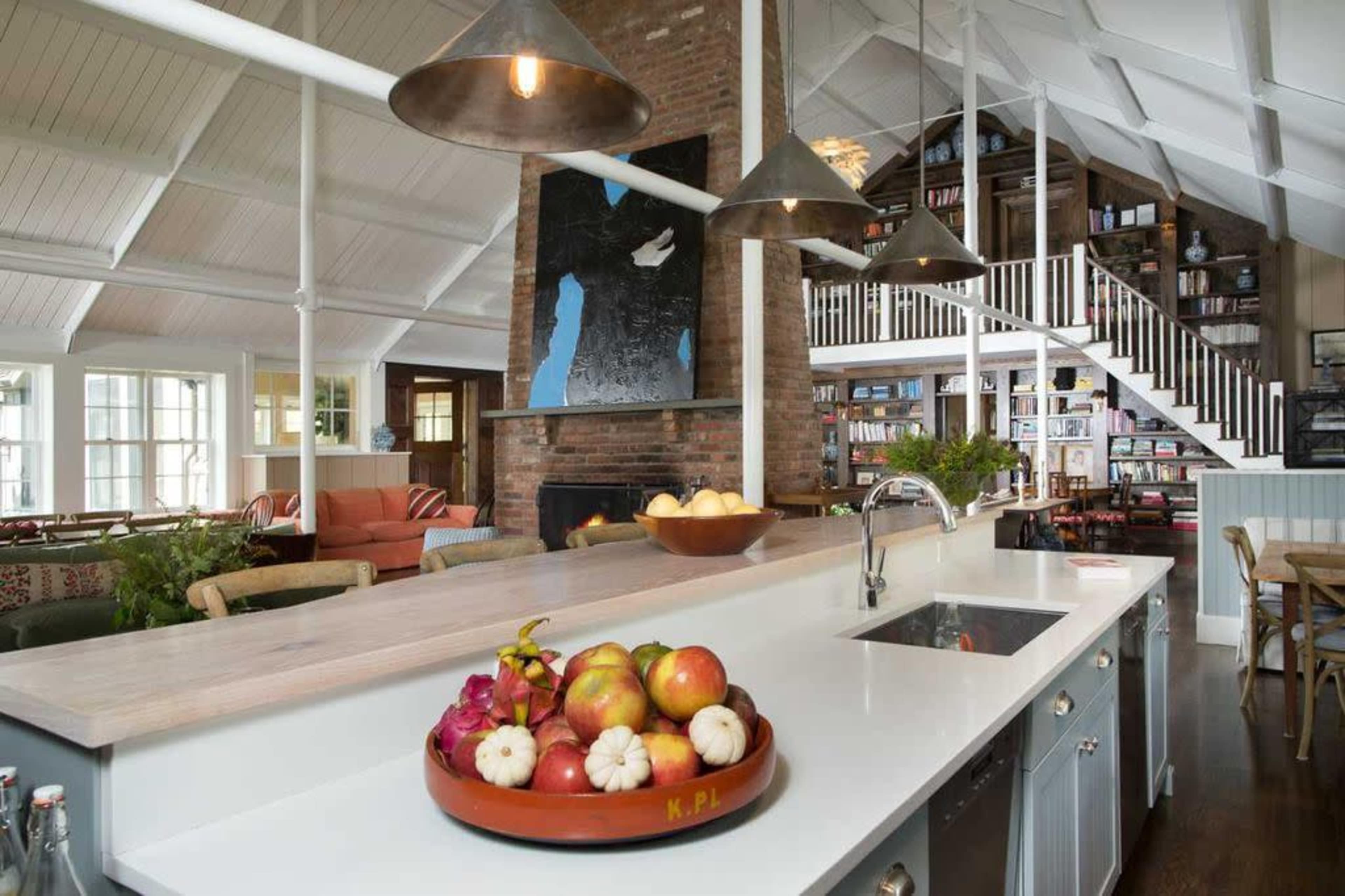 The image shows a spacious kitchen with a central island, featuring a bowl of colorful fruits, and an open living area connected to a staircase leading to a library.