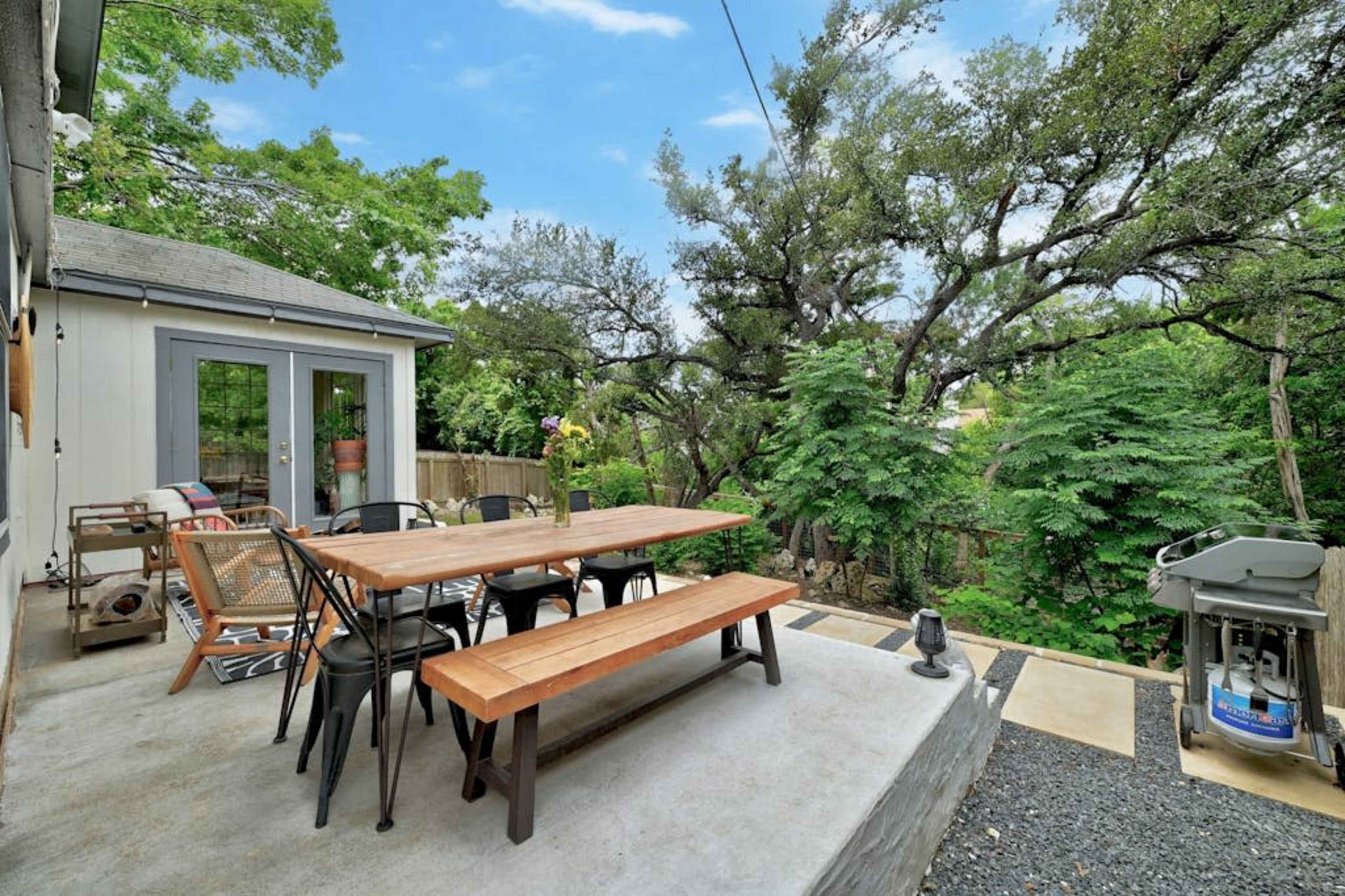 The image shows a patio area with a wooden dining table and chairs surrounded by lush greenery and trees.