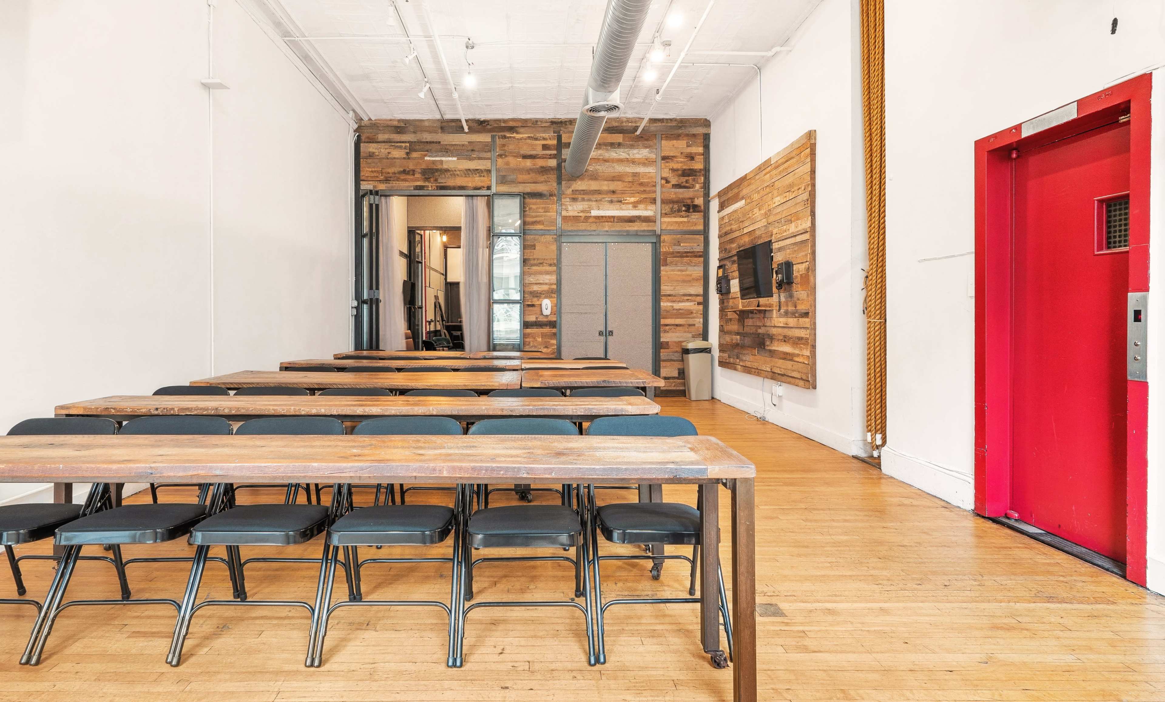 A spacious room with wooden tables and chairs arranged in a row, featuring a red elevator on the right and a wood-paneled wall on the left.