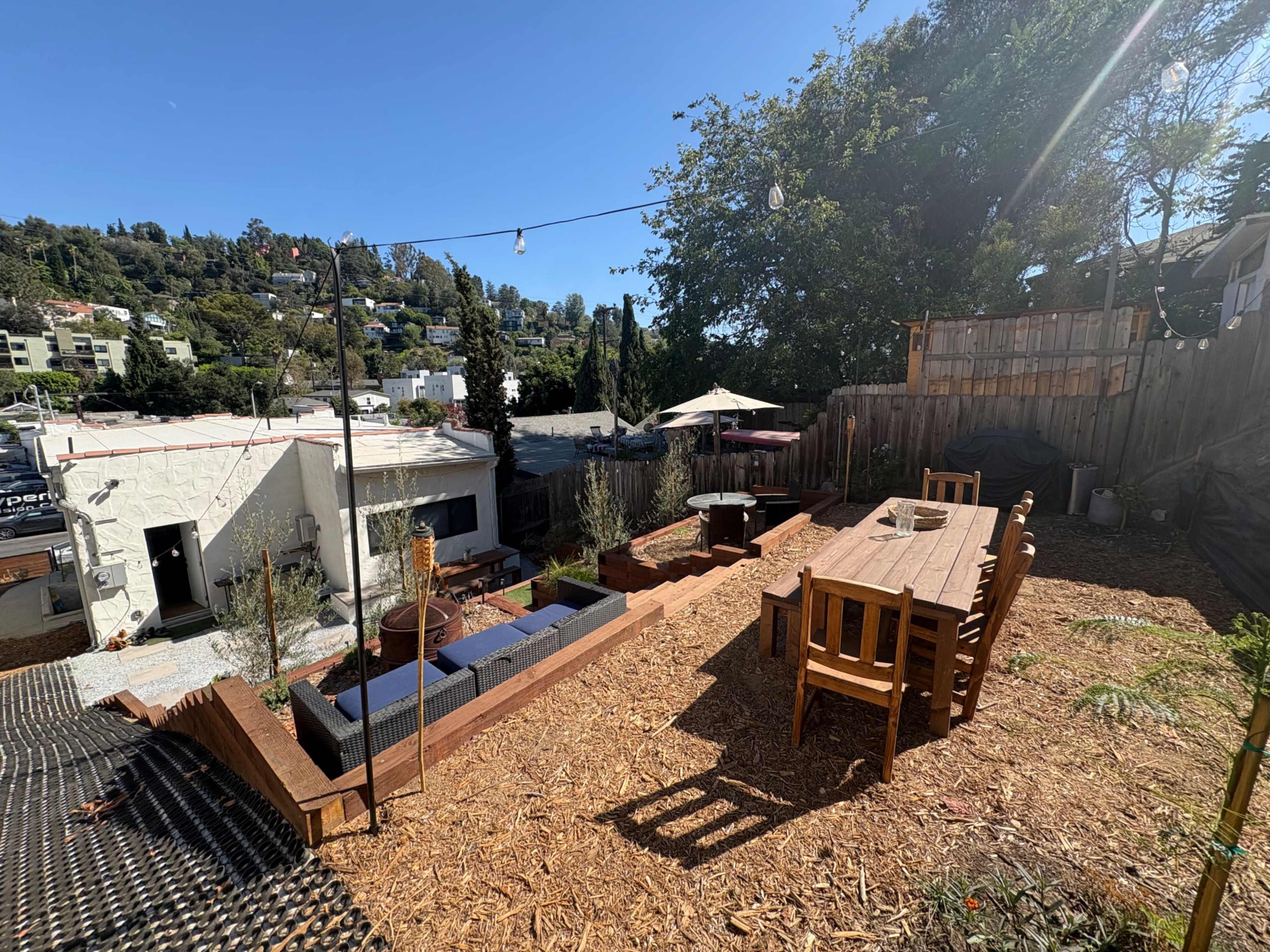 The image shows a landscaped backyard featuring a wooden dining table with chairs, garden beds, a small seating area, and a view of surrounding houses and trees.