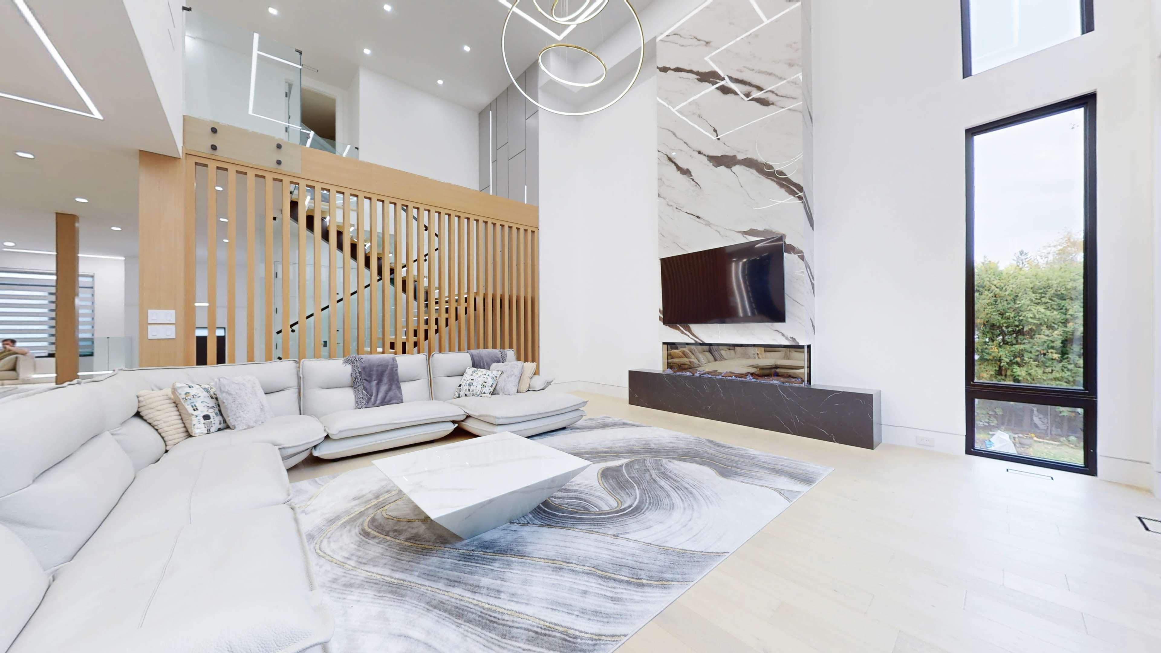 A modern living room featuring a large sectional sofa, a marble coffee table, and a wall-mounted television above a marble fireplace, all set within a bright space characterized by high ceilings and wooden accents.