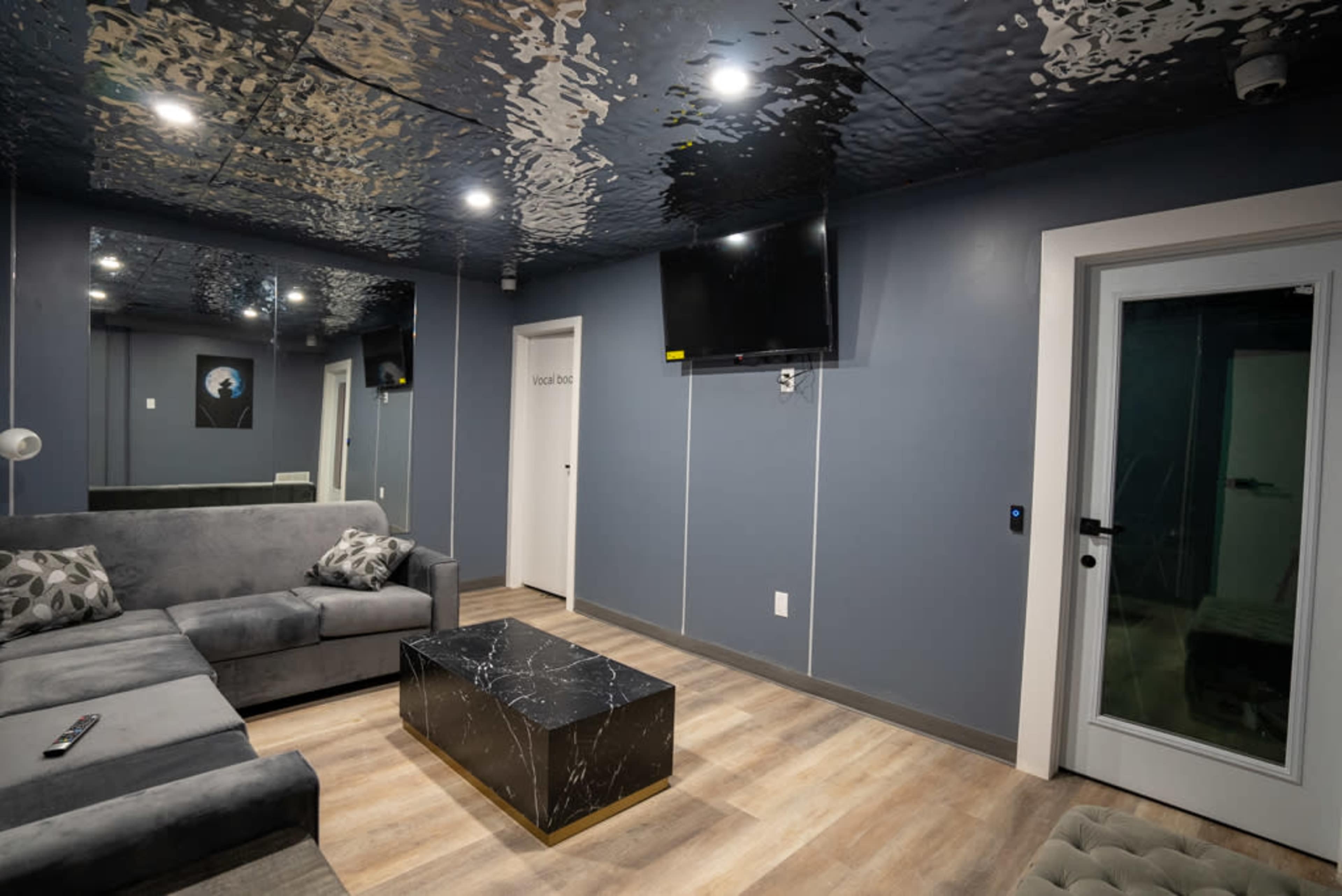 A modern lounge area features a gray sectional sofa, a black marble coffee table, a flat-screen TV, and a door leading to another room, all under a reflective ceiling.