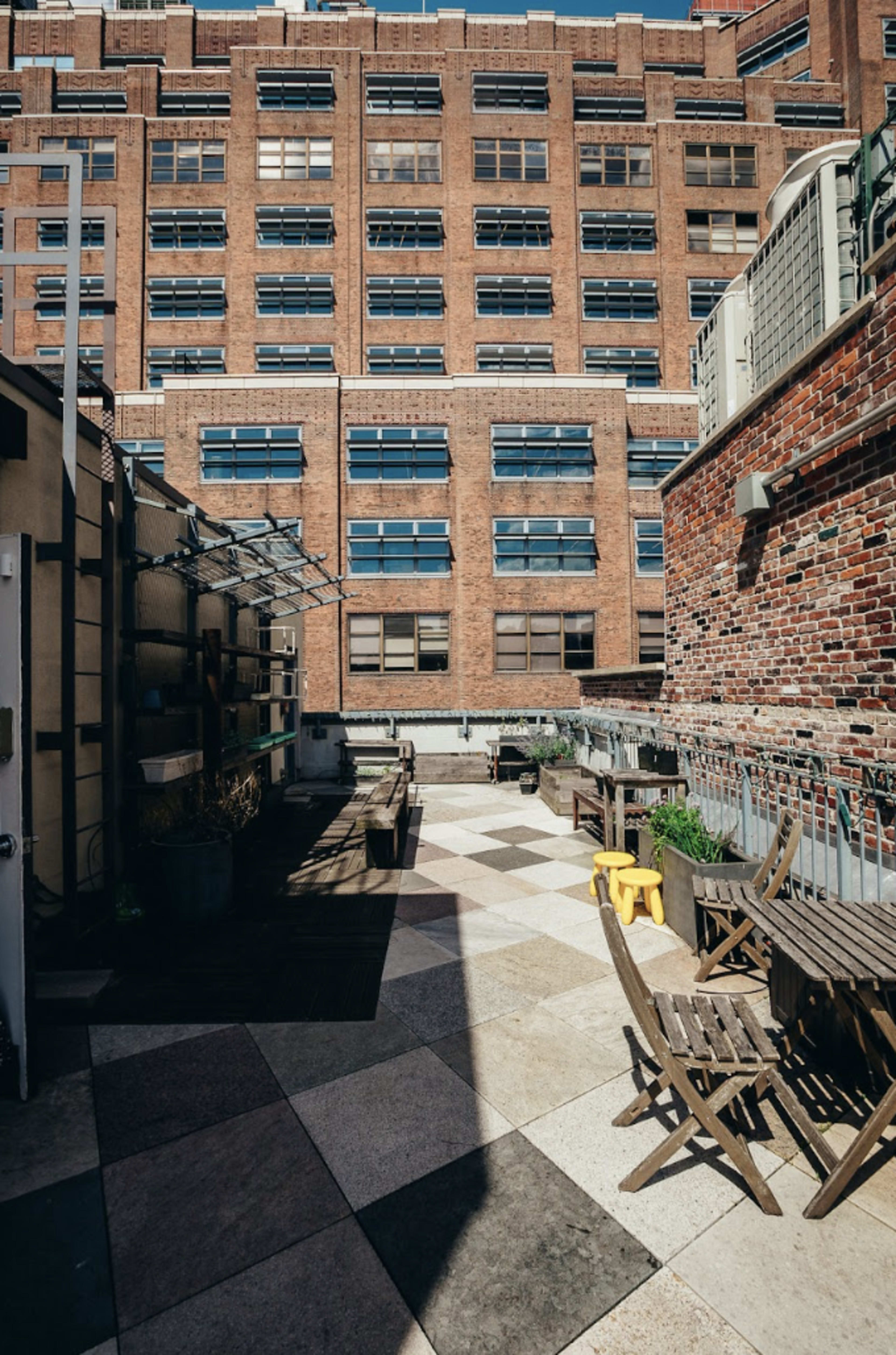 The image shows a rooftop terrace featuring various wooden furniture, potted plants, and a view of a brick building with multiple windows in the background.