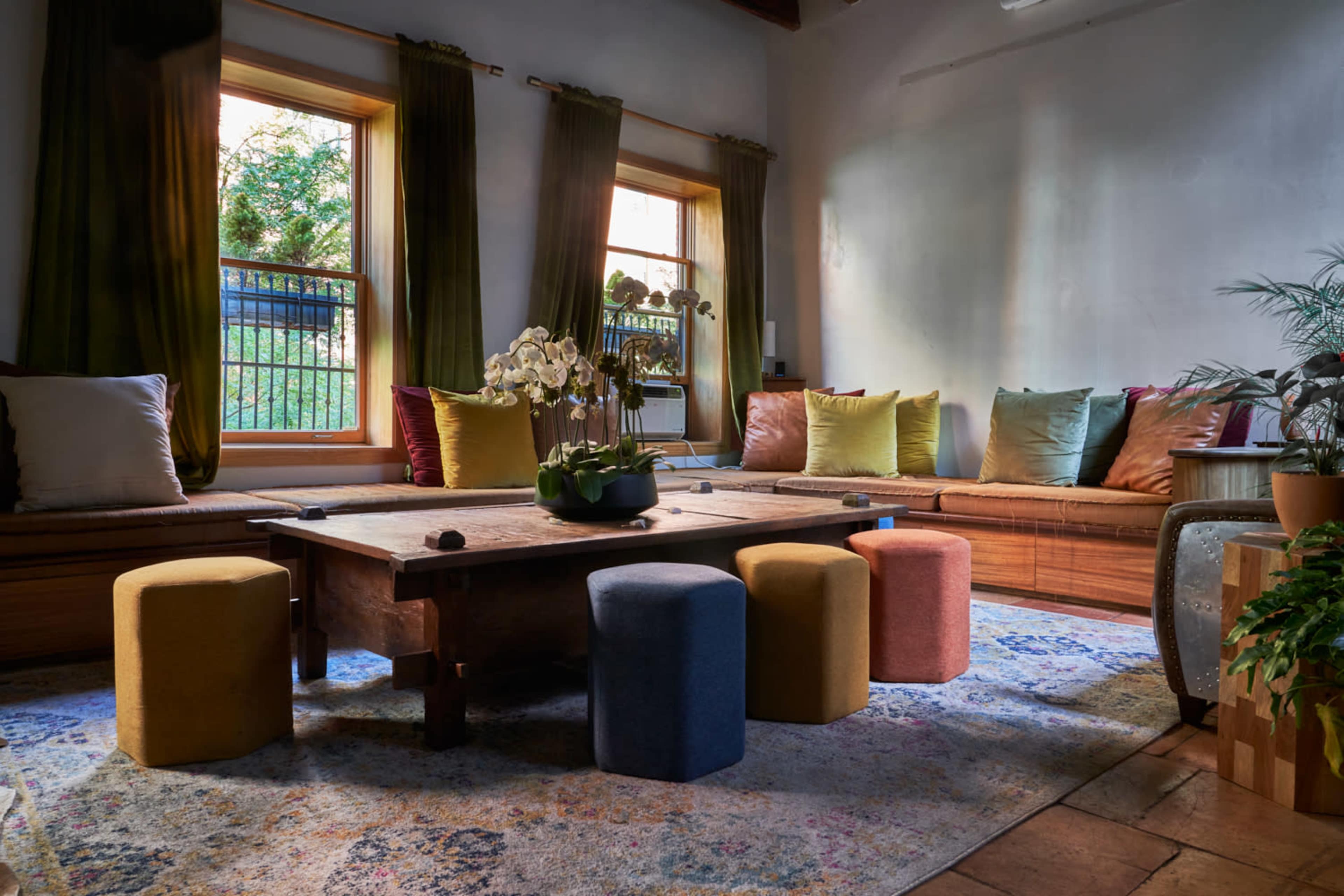 A cozy room with a wooden table surrounded by colorful stools and a seating area adorned with cushions near large windows.