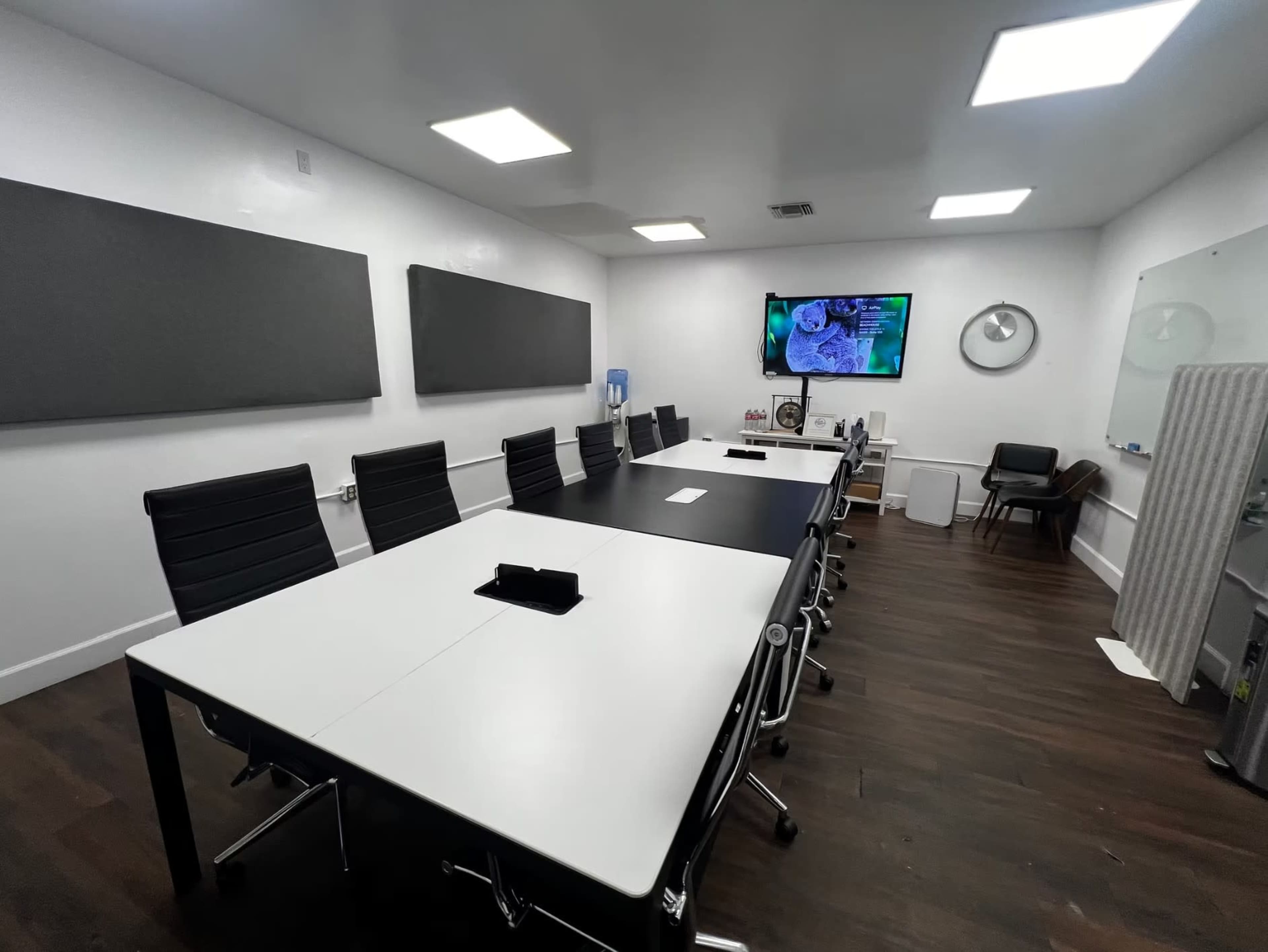The image shows a modern conference room equipped with a long table, black and white seating, wall-mounted screen, and soundproof panels.