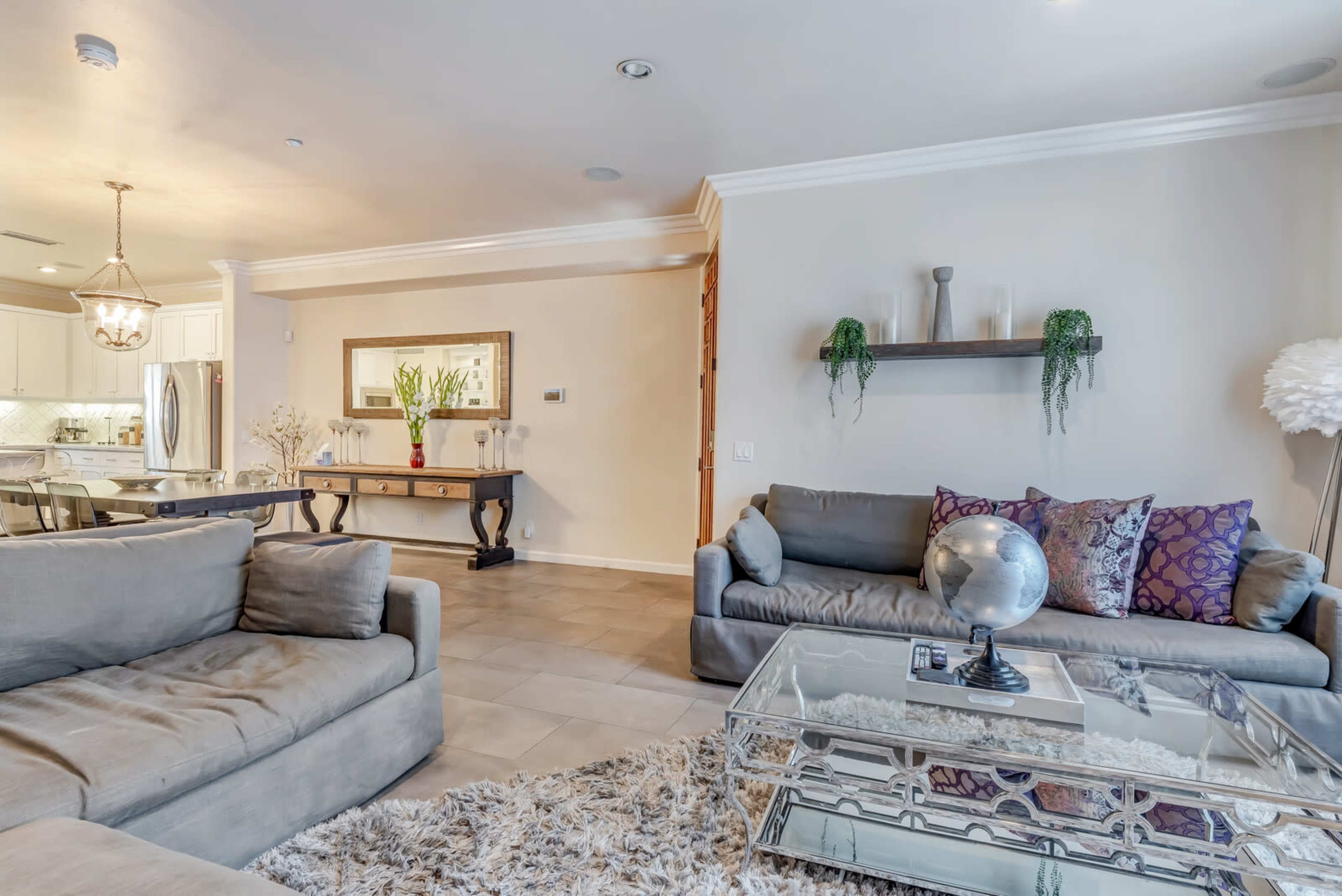 A contemporary living room features two gray sofas, a glass coffee table, and a decorative mirror on the wall, with adjacent kitchen visible in the background.