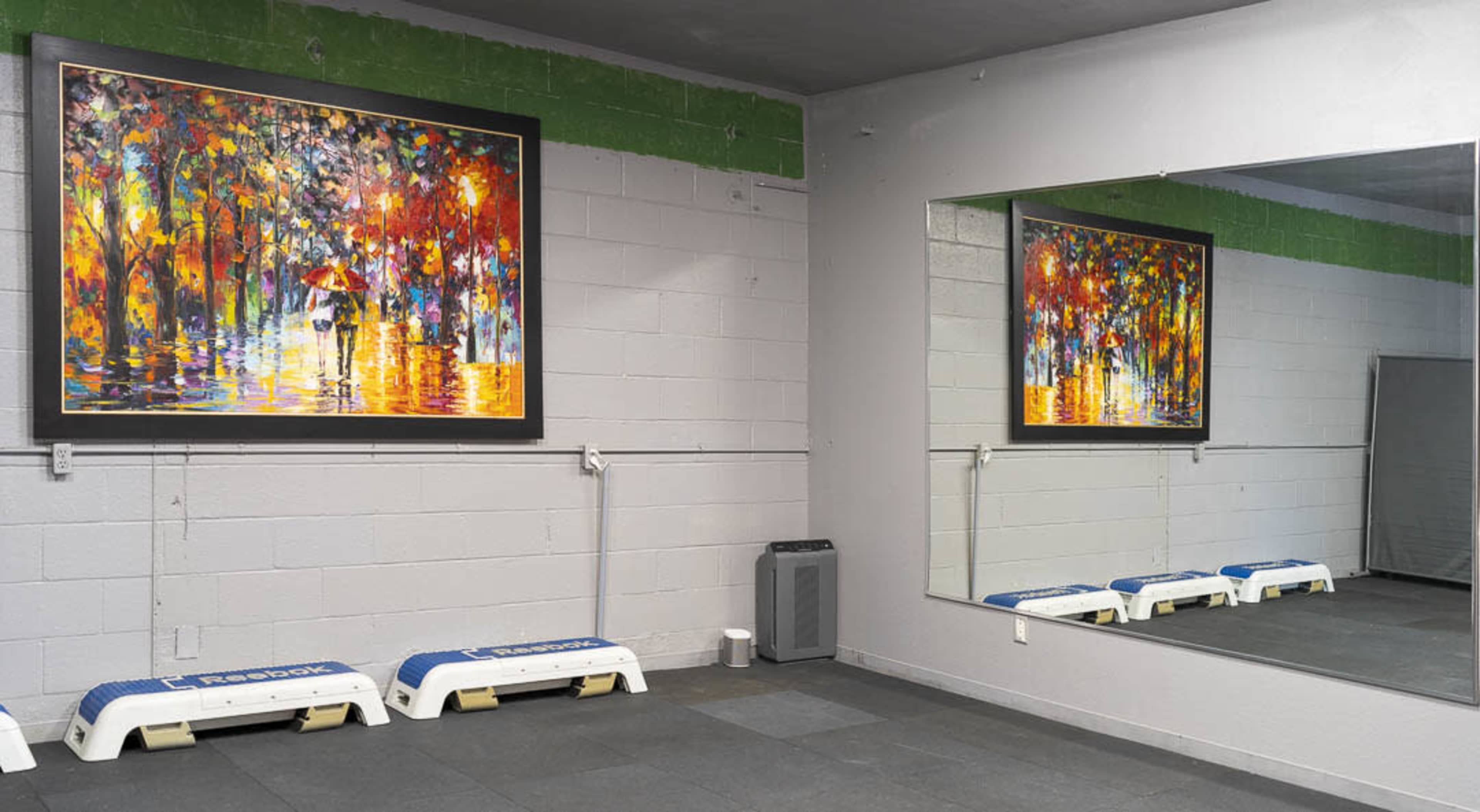 A gym area with a large colorful painting on the wall, a mirror reflecting the painting, and two white exercise benches positioned on the floor.