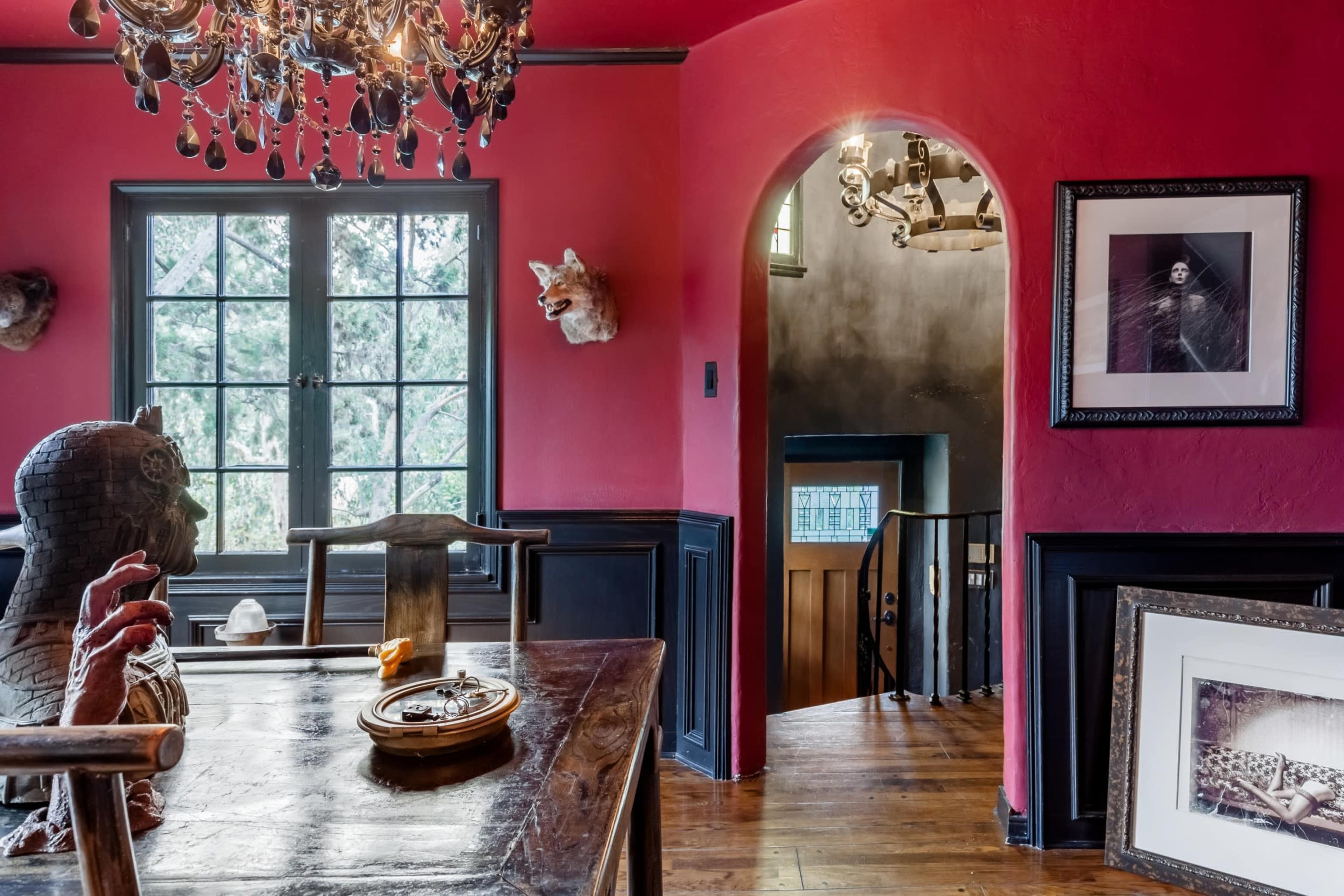 A dining area with a dark wooden table, a large chandelier, red-painted walls, and an arched doorway leading to a stairwell.