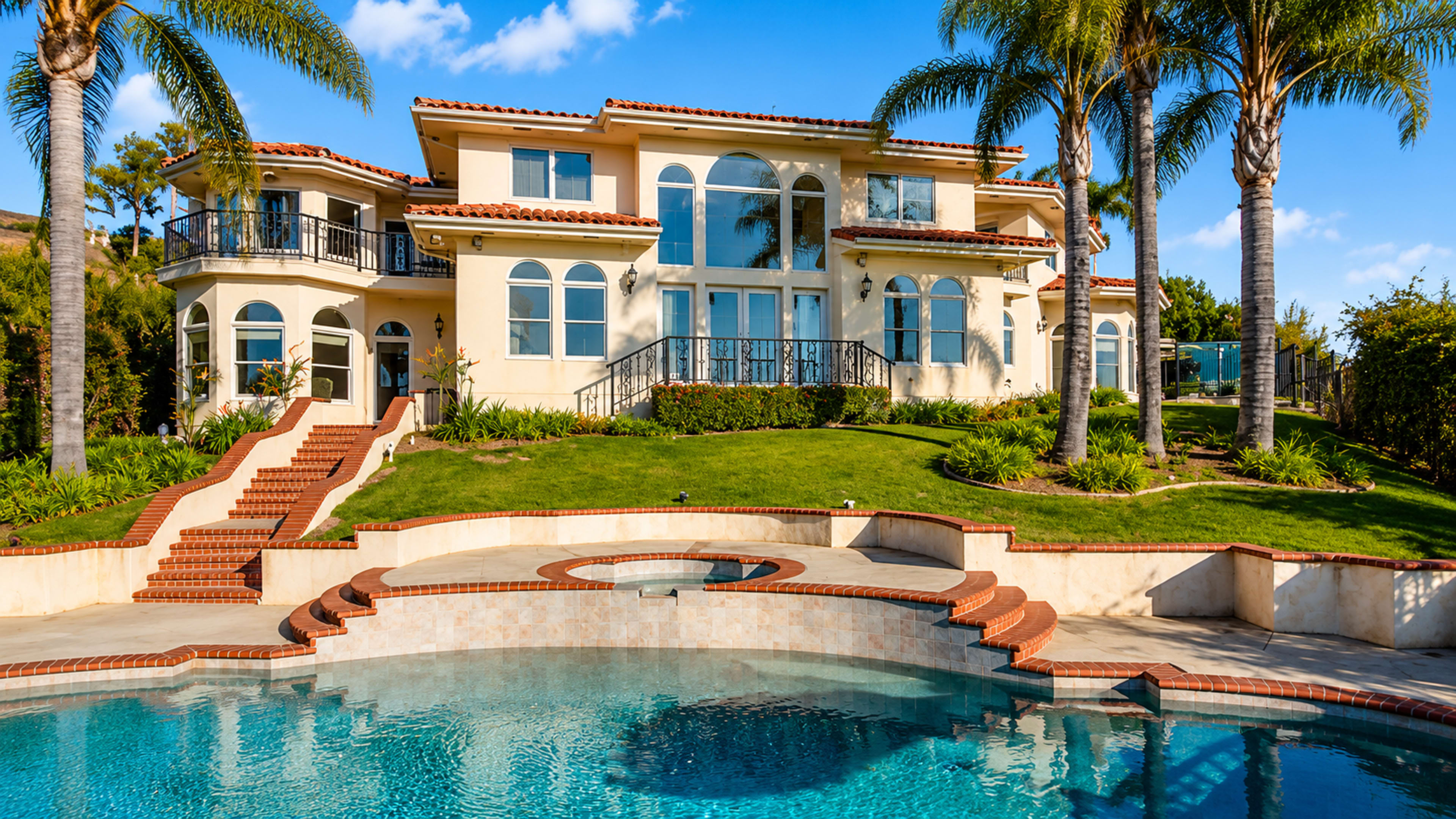 A large, luxurious house features multiple balconies, large windows, and is surrounded by palm trees and a well-maintained lawn, with a swimming pool in the foreground.