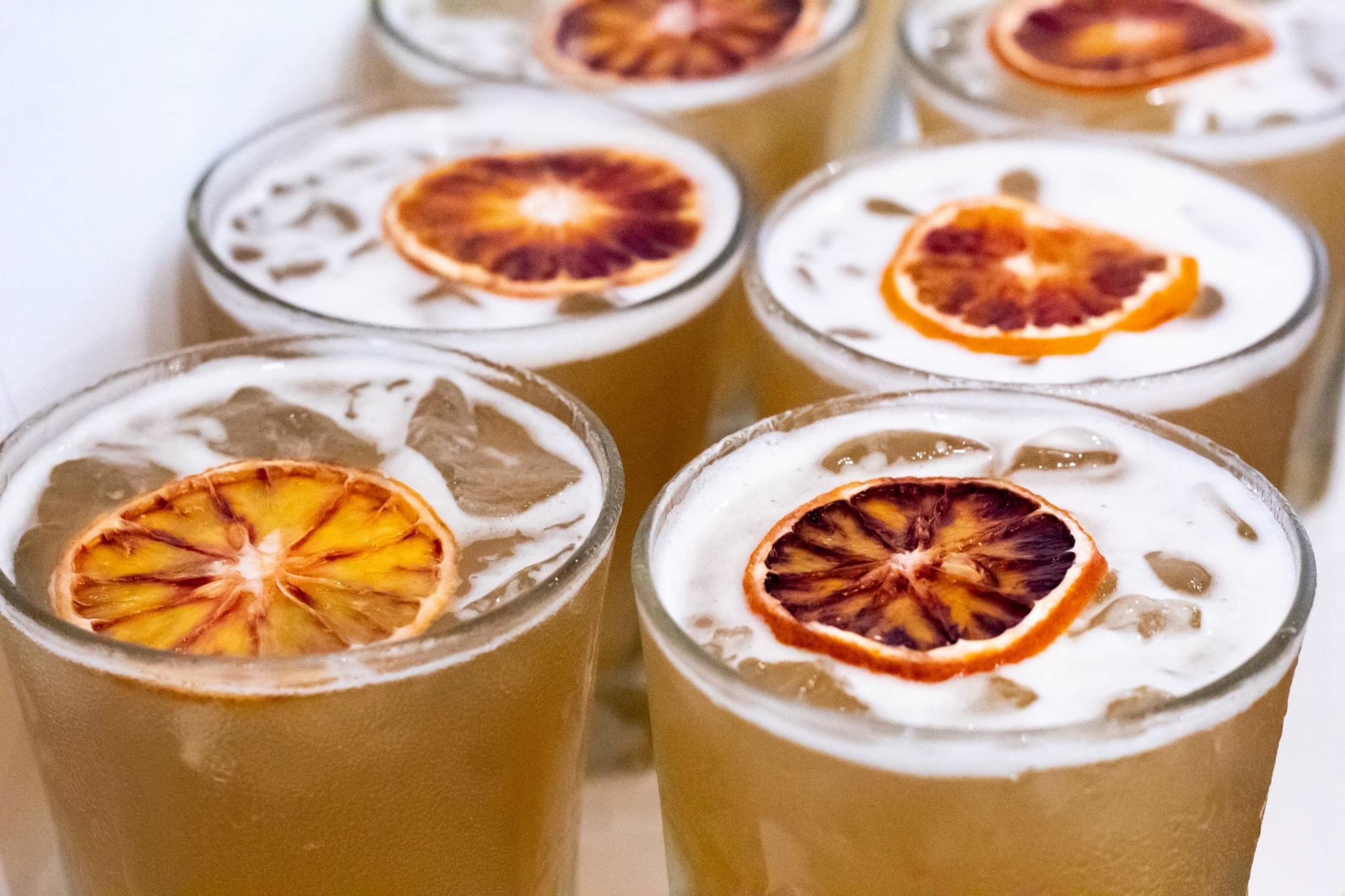 Several glasses filled with a light-colored beverage and topped with ice and dried orange slices are arranged closely together.